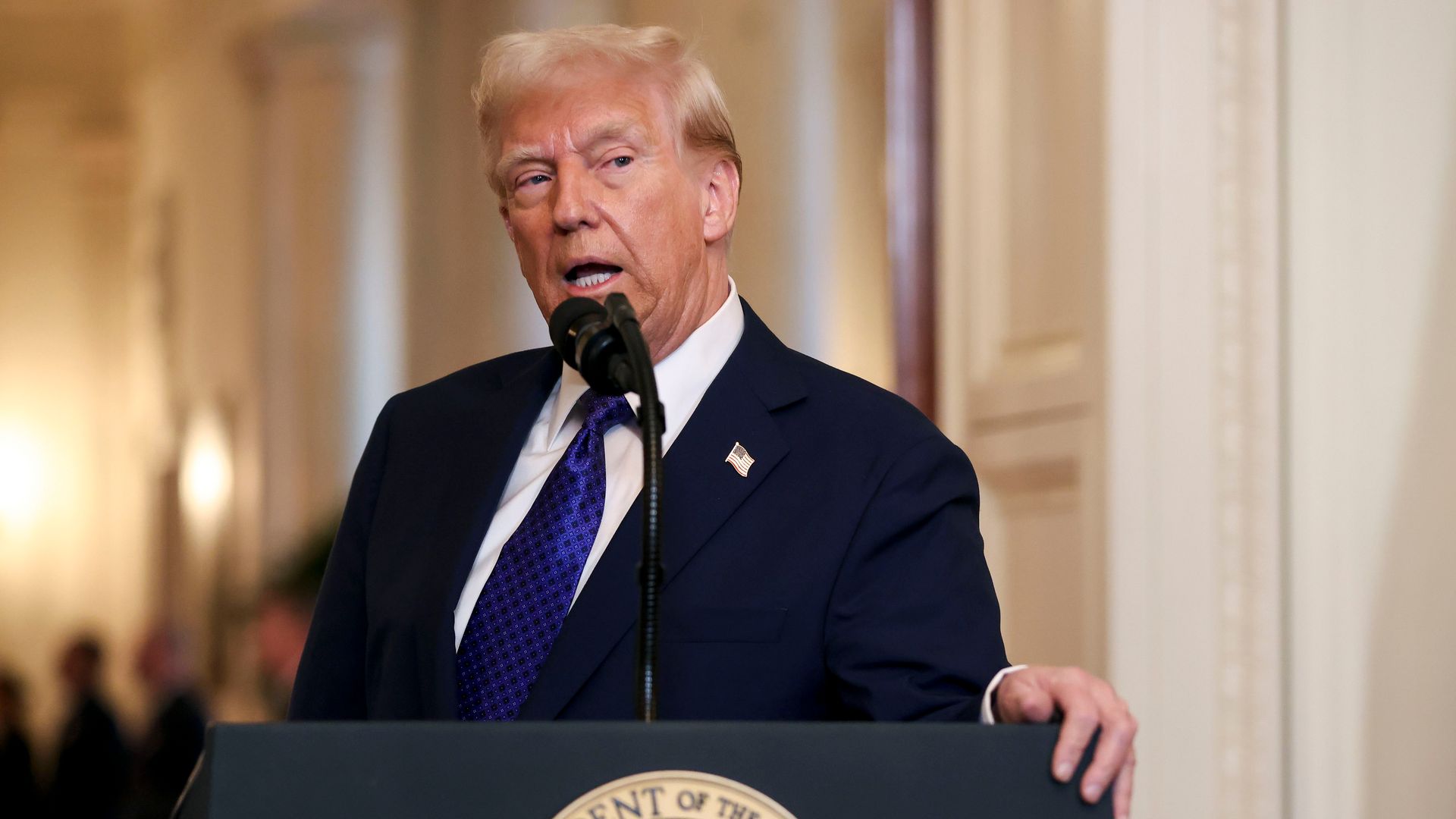 Donald Trump, wearing a blue suit and gripping a podium in a white room.