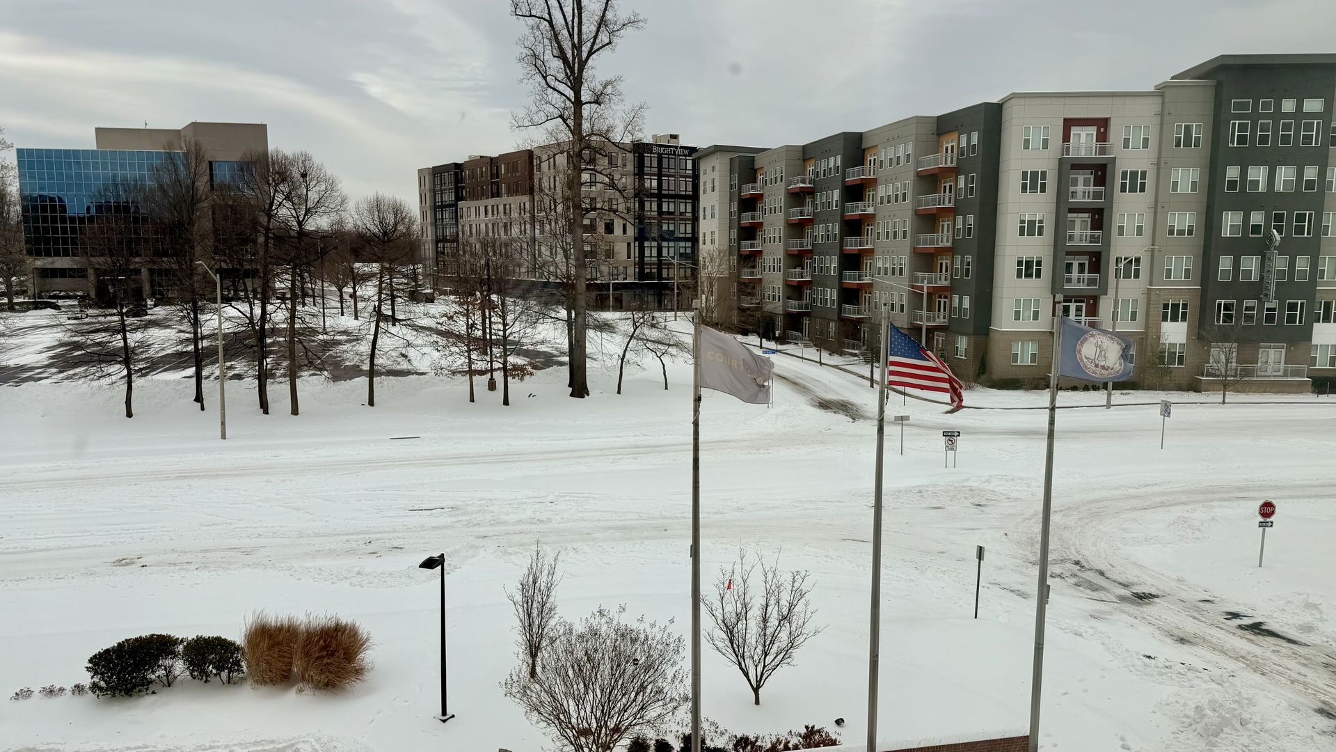 Snow-covered street and sidewalks in front of modern apartment and office buildings on a gray winter day, with bare trees, three flagpoles flying American and other flags.