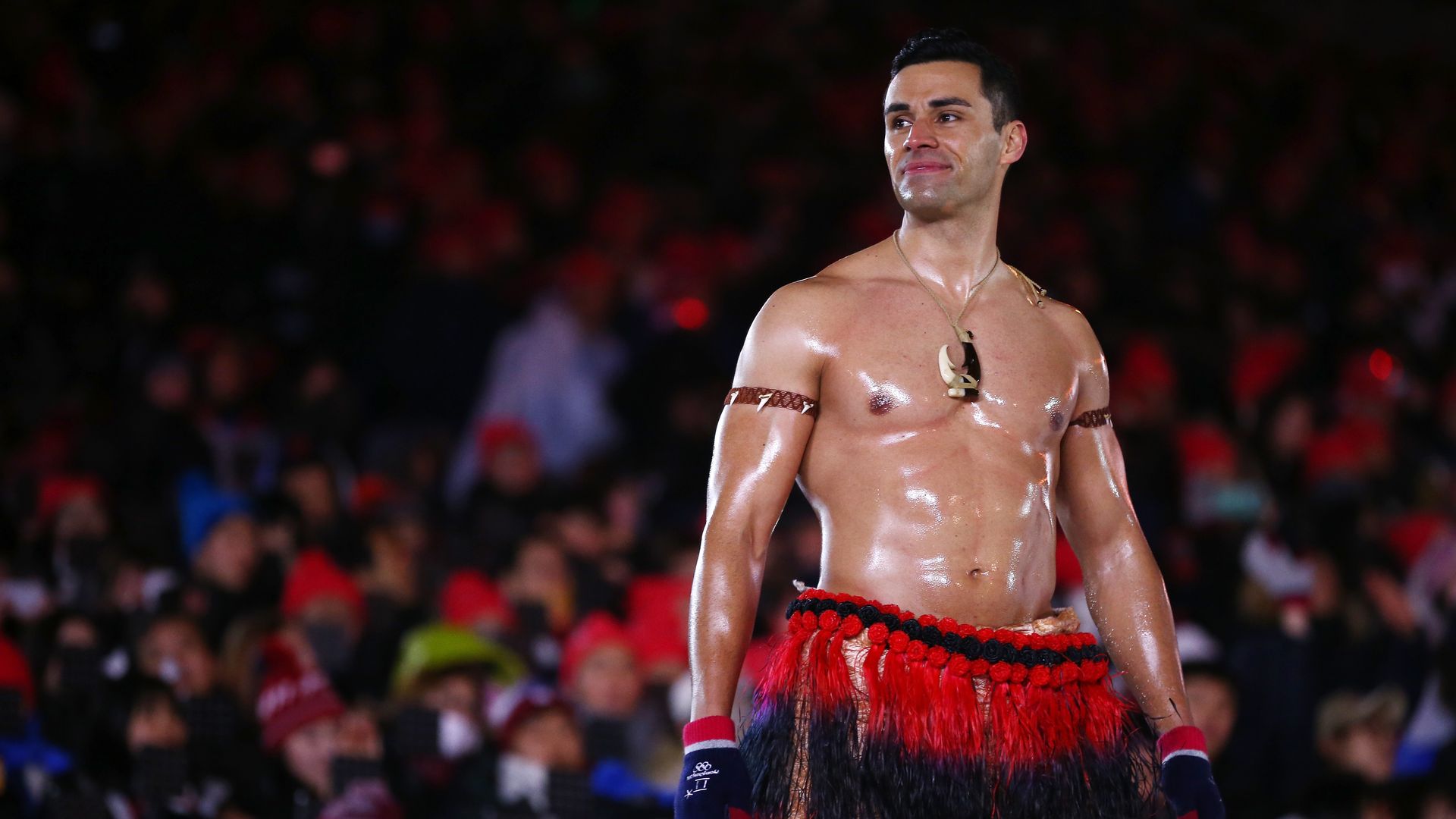Photo of a shirtless, oiled up Pita Taufatofua wearing red and black traditional clothing on the bottom and smiling at the crowd