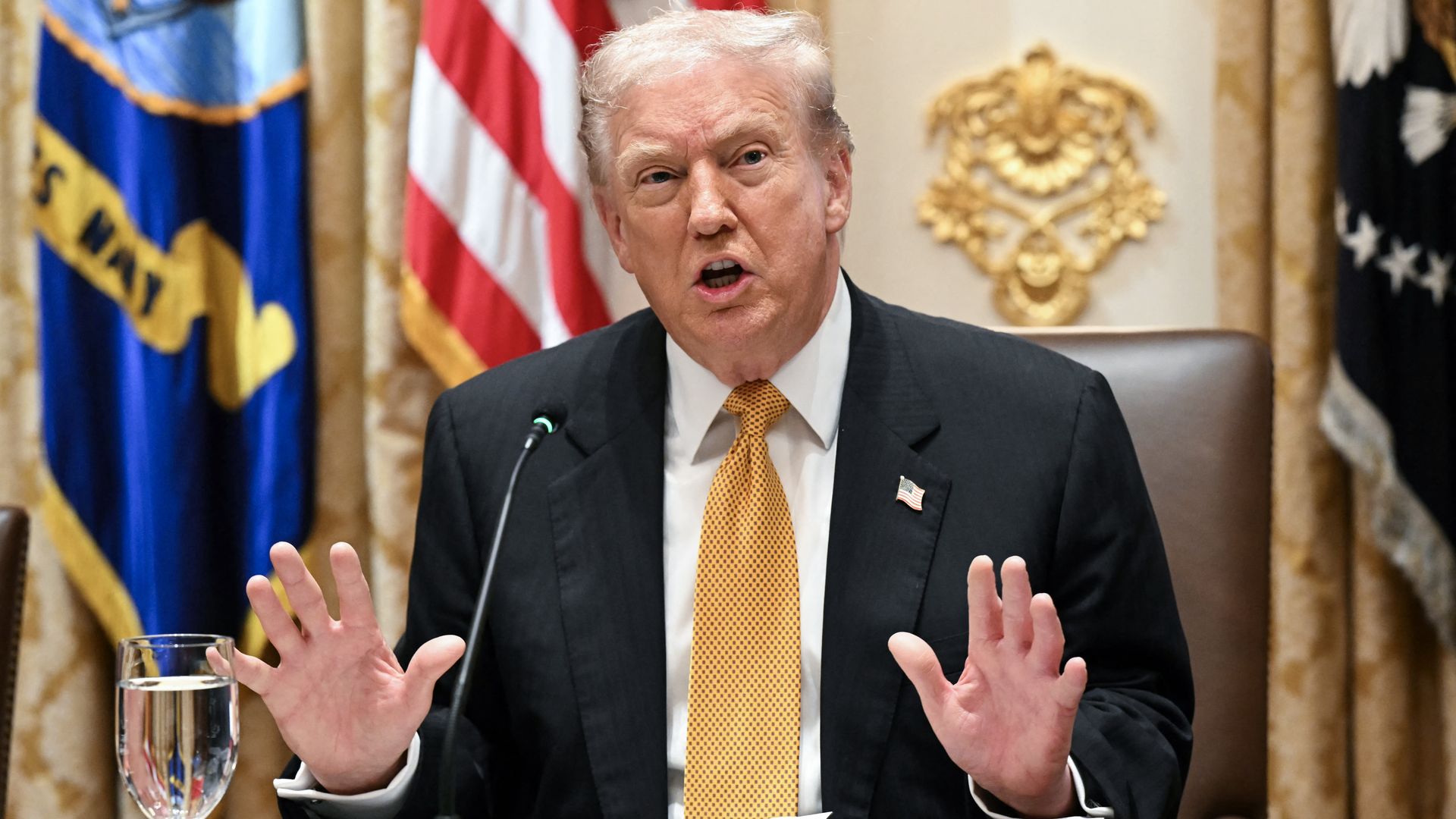 President Trump, in a black suit and gold tie, gestures with both hands as he sits in front of a series of flags. 