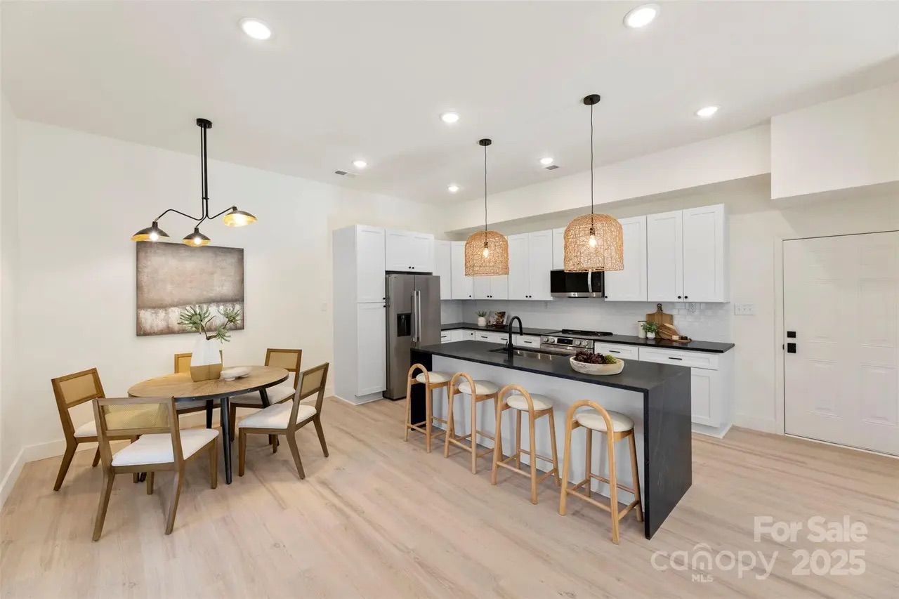 Bright modern kitchen with white cabinets, black countertops, stainless steel appliances, four wooden bar stools at the island, and a round dining table with five wooden chairs.