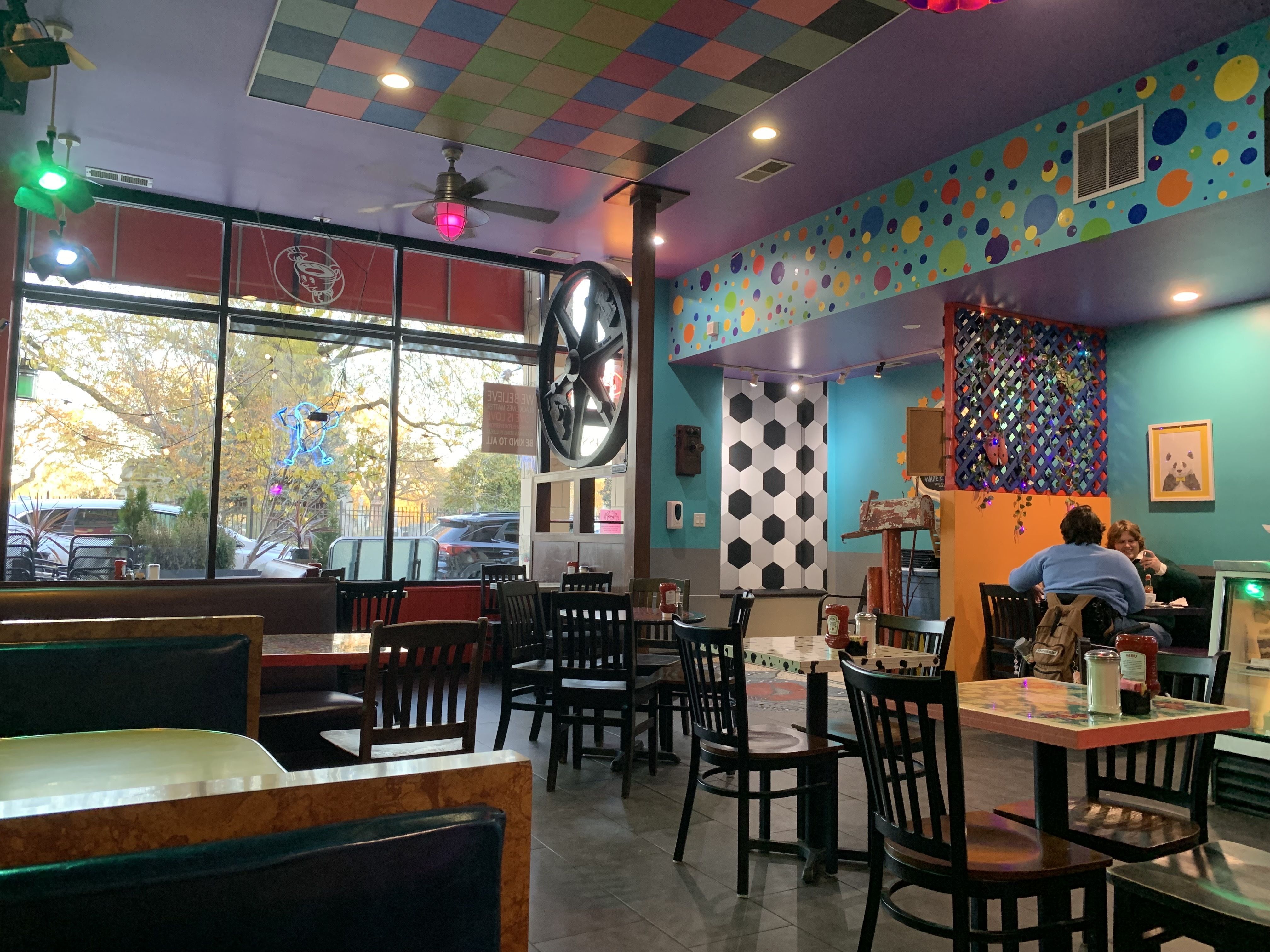 Colorful café interior with teal and purple walls, a multicolored checkered ceiling, patterned furniture, and a large window showing trees and parked cars outside.