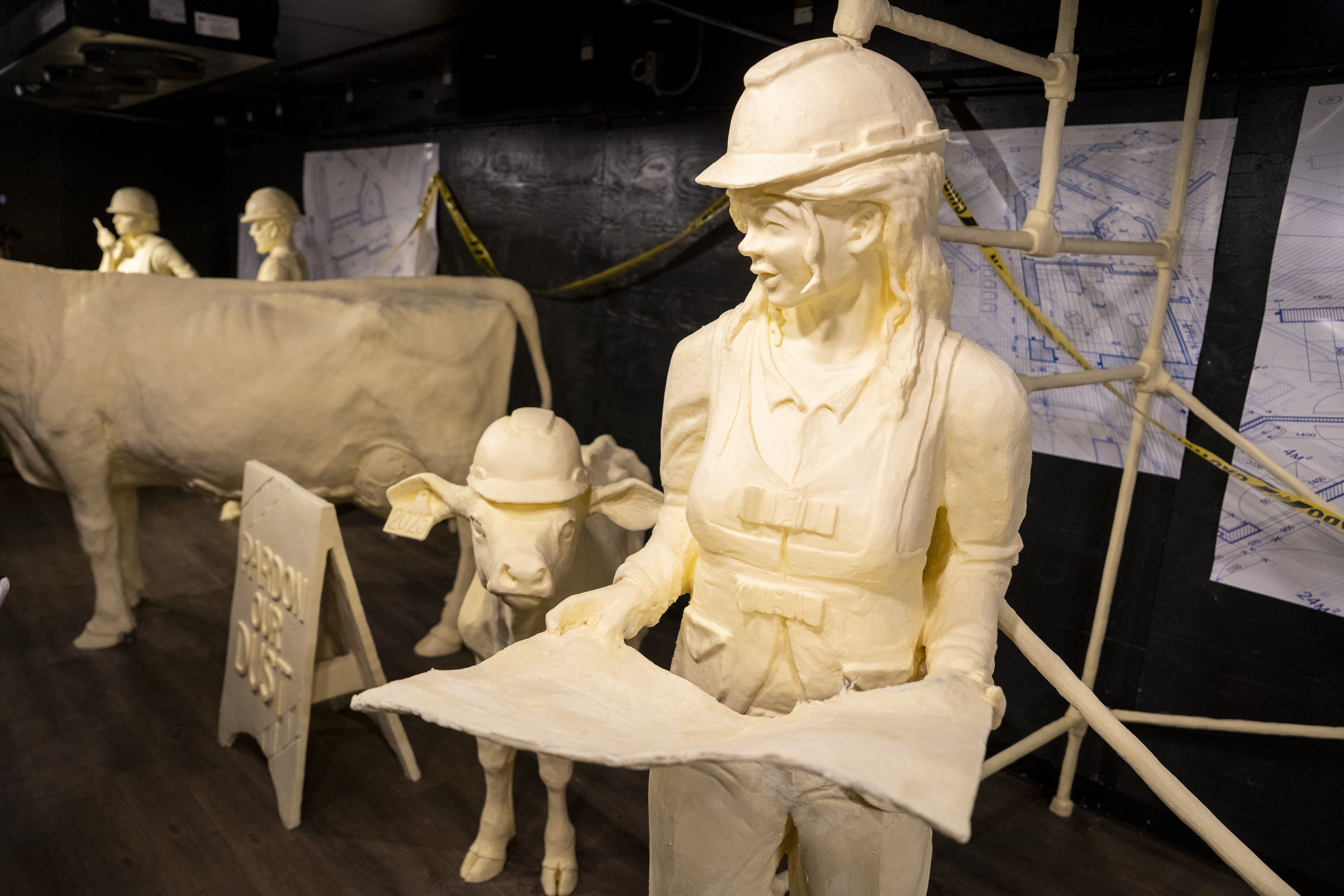 Sculptures of a female construction worker holding blueprints, a calf wearing a hard hat, and a cow, all made from a pale material, displayed with construction blueprints and caution tape.