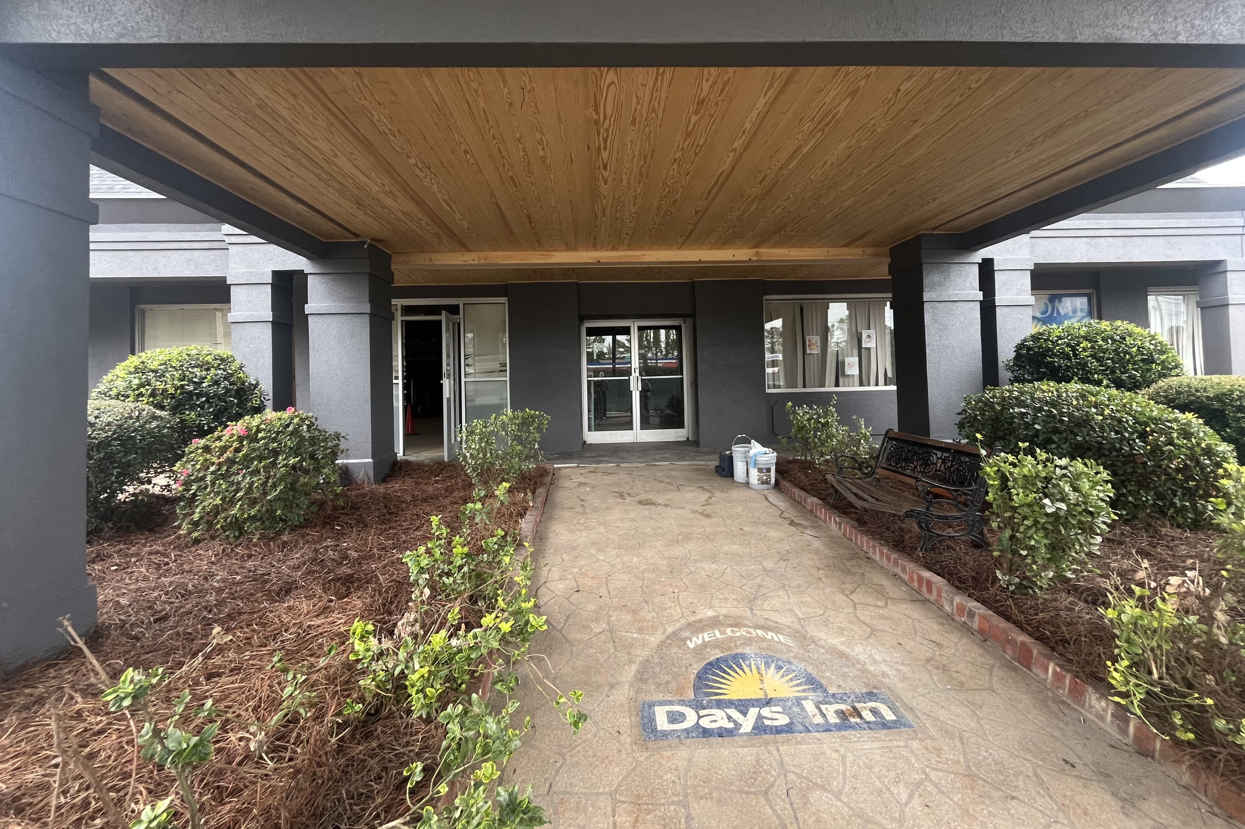 Covered entrance of a hotel with gray pillars, wooden ceiling, bushes on either side, a bench, and the Days Inn logo painted on the ground near double glass doors.
