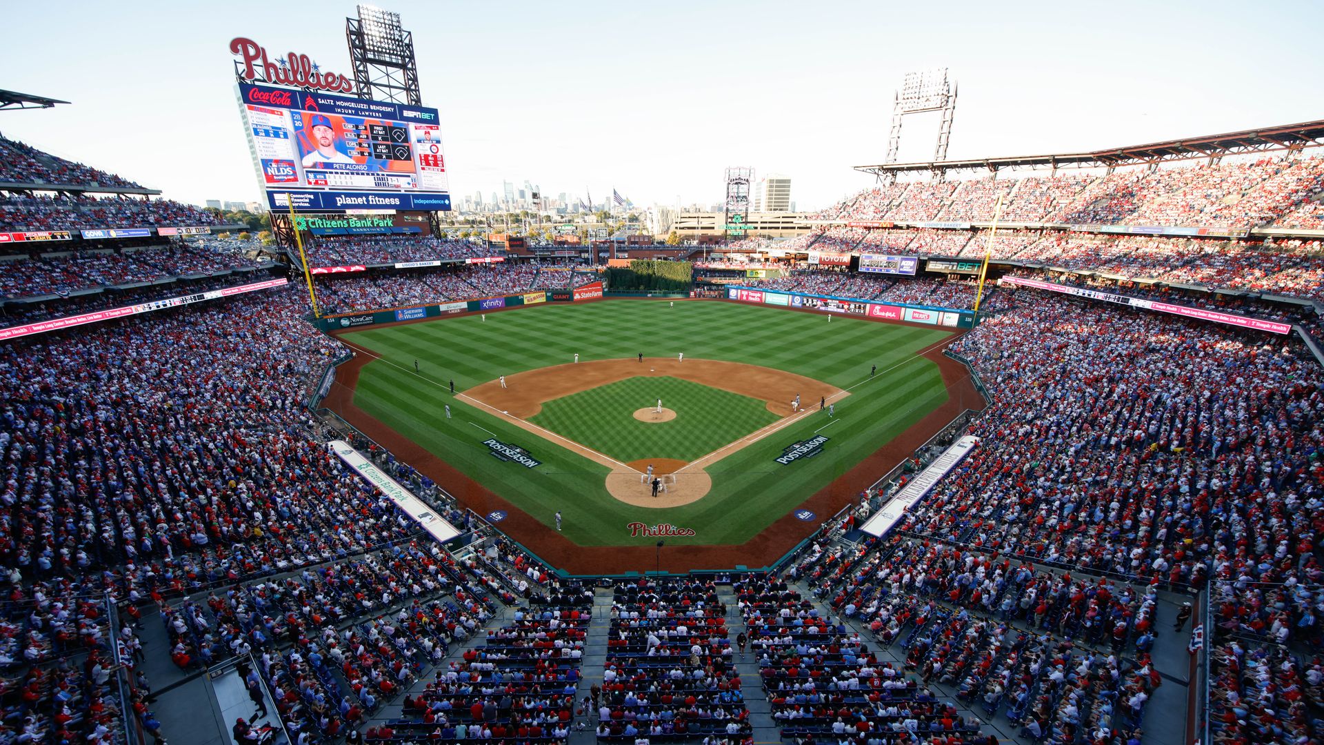 Full baseball stadium packed with fans for a day game. Green outfield, dirt infield, players on the diamond, a large Phillies scoreboard, and a distant city skyline.