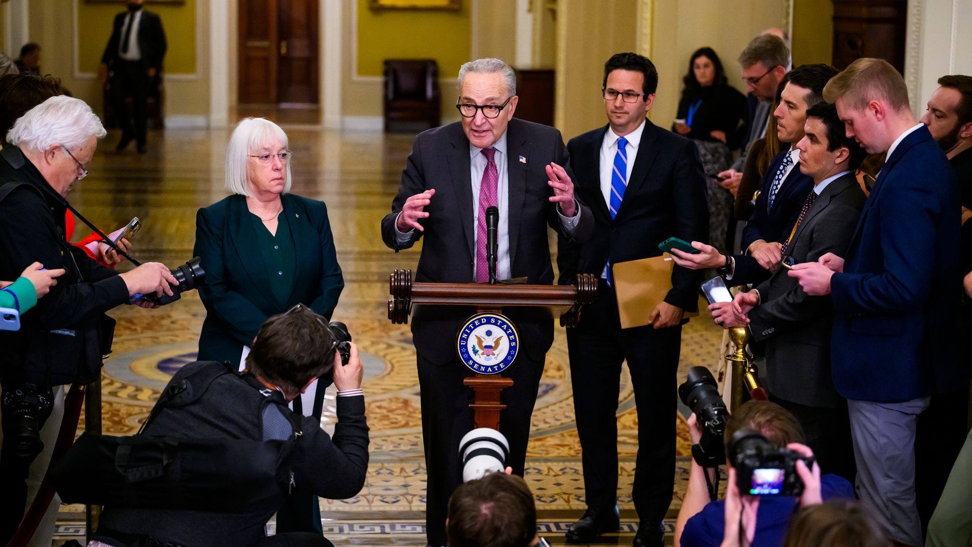 Sens. Patty Murray, Chuck Schumer and Brian Schatz speak to a throng of reporters and photographers in a yellow hallway with an ornate tiled floor.