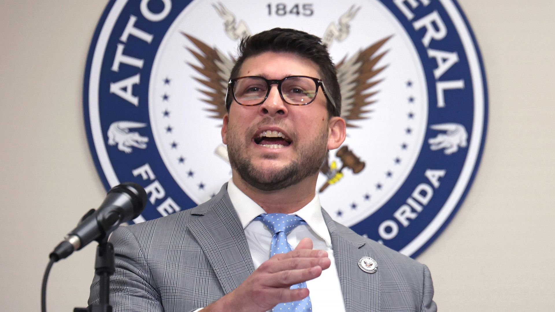 Man with glasses speaking at a podium with a microphone, wearing a gray checked suit and light blue tie, in front of a circular 'Attorney General Florida' seal.
