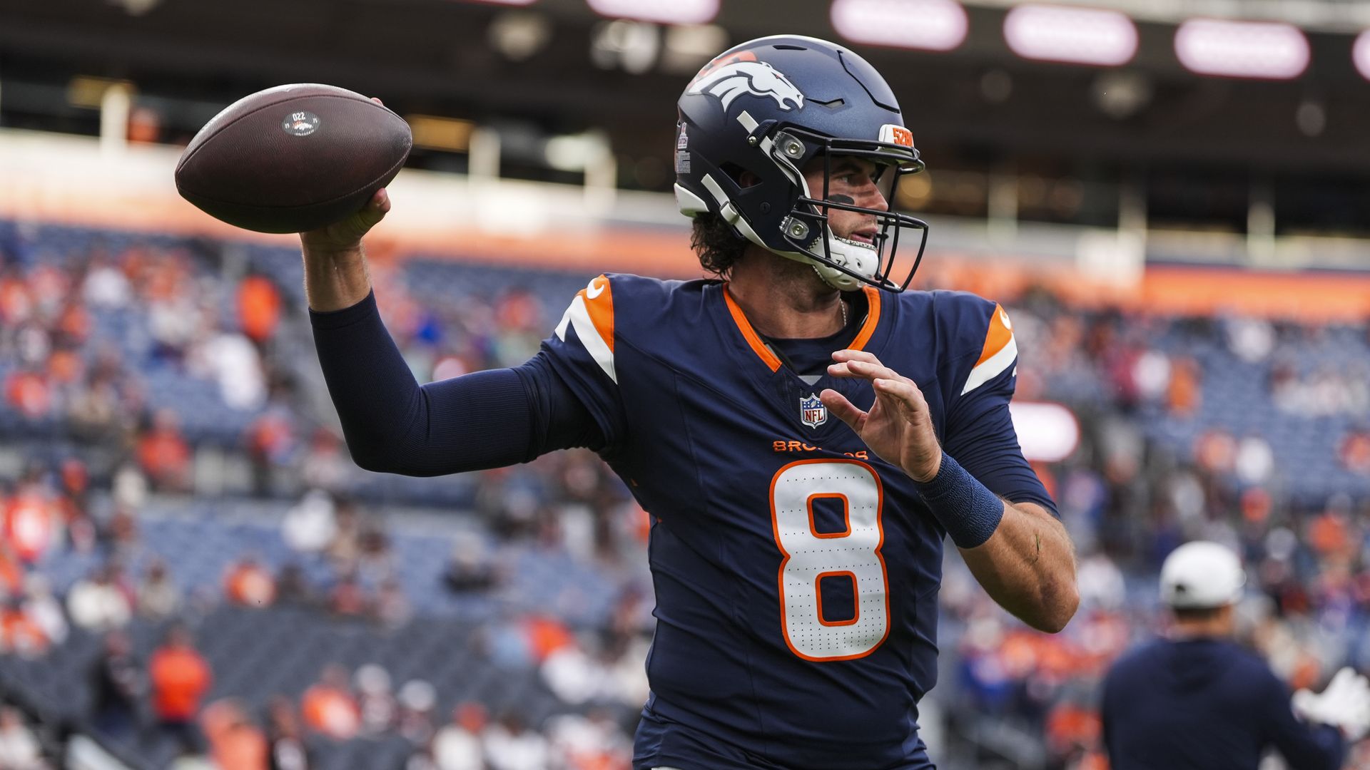 Denver Broncos football player wearing navy blue and orange uniform with number 8 throwing a football in a stadium with blurred spectators in the background.