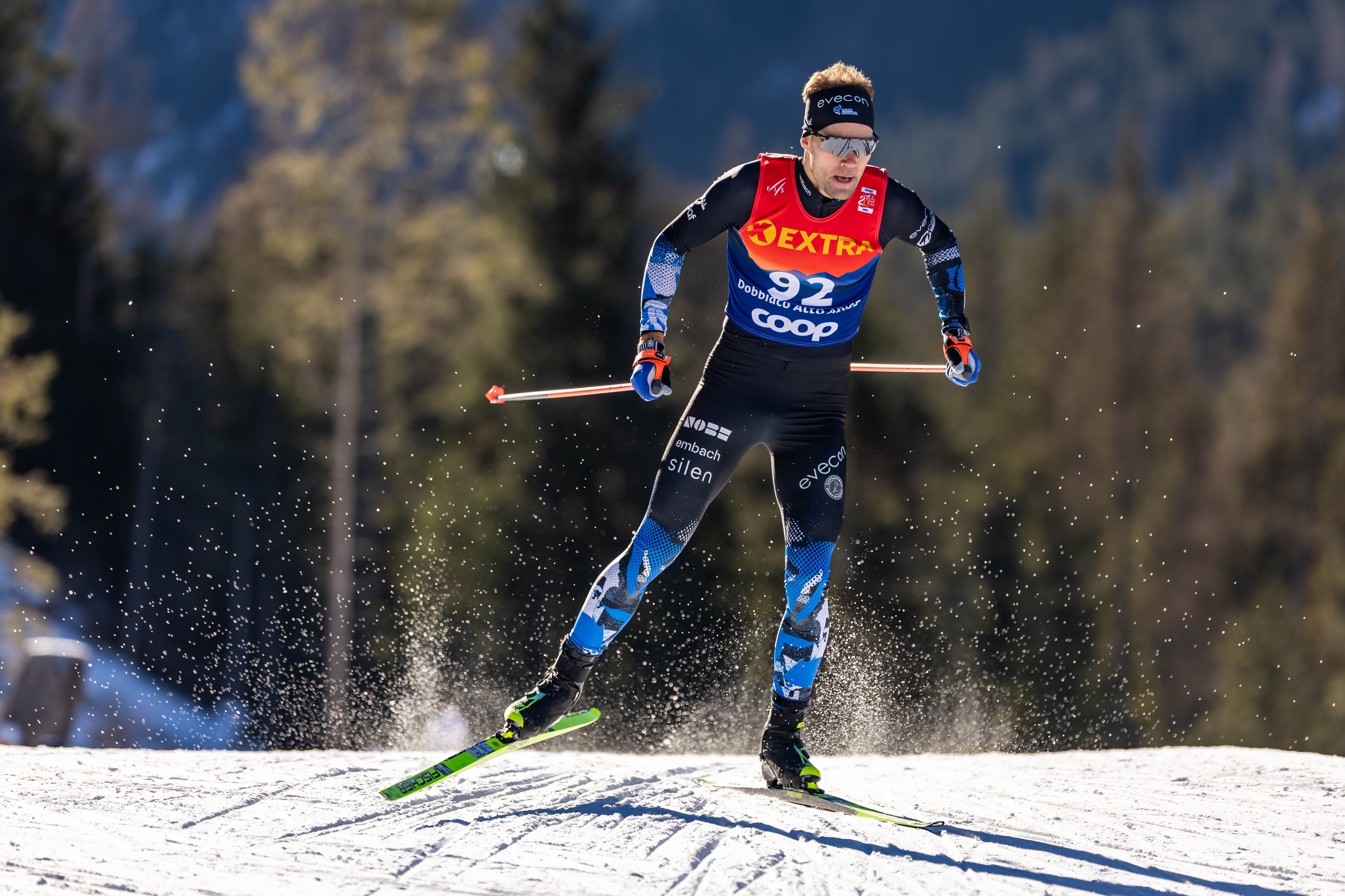 Cross-country skier wearing black, blue, and red with bib number 92 racing on snowy trail, using ski poles, with forest and blue sky in background.