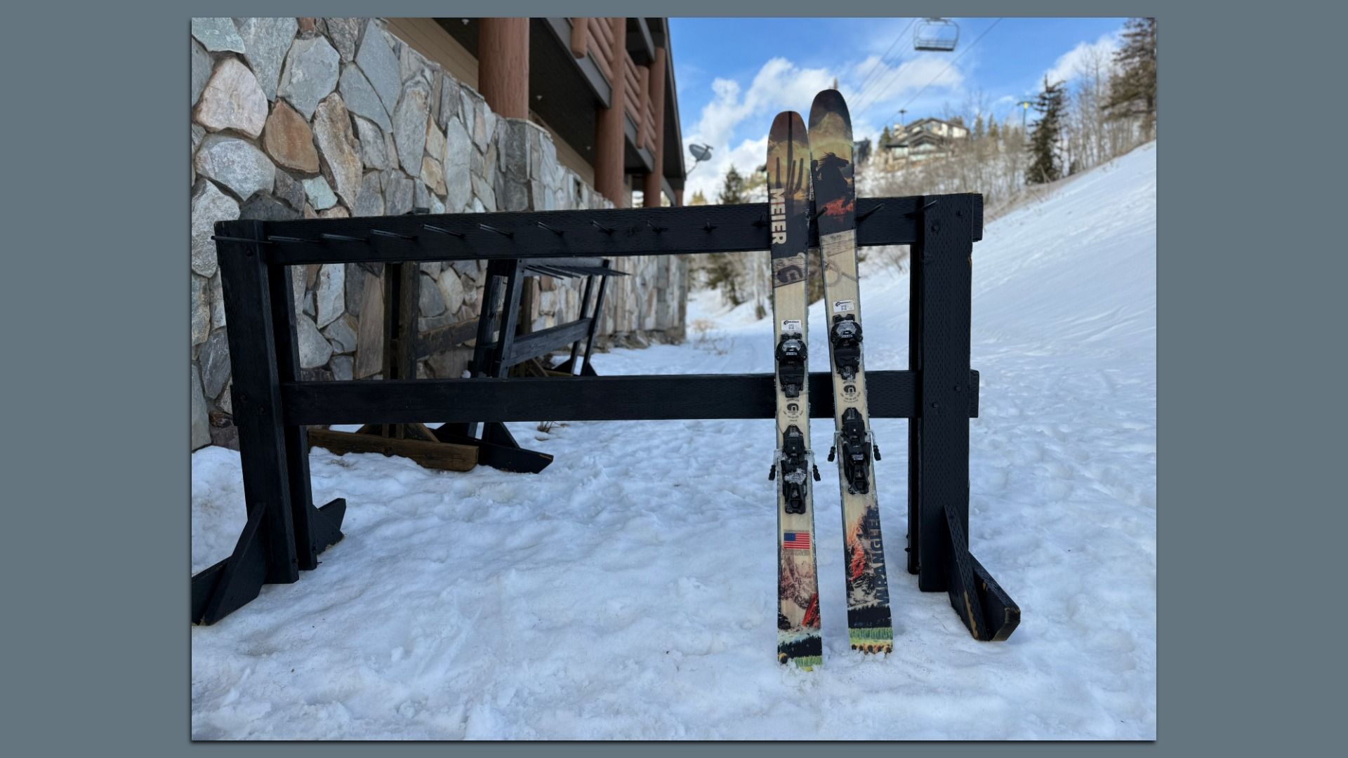 Two alpine skis with bindings leaned in a black wooden ski rack on a snow-covered slope, beside a stone wall and a lodge, under a blue sky with clouds.