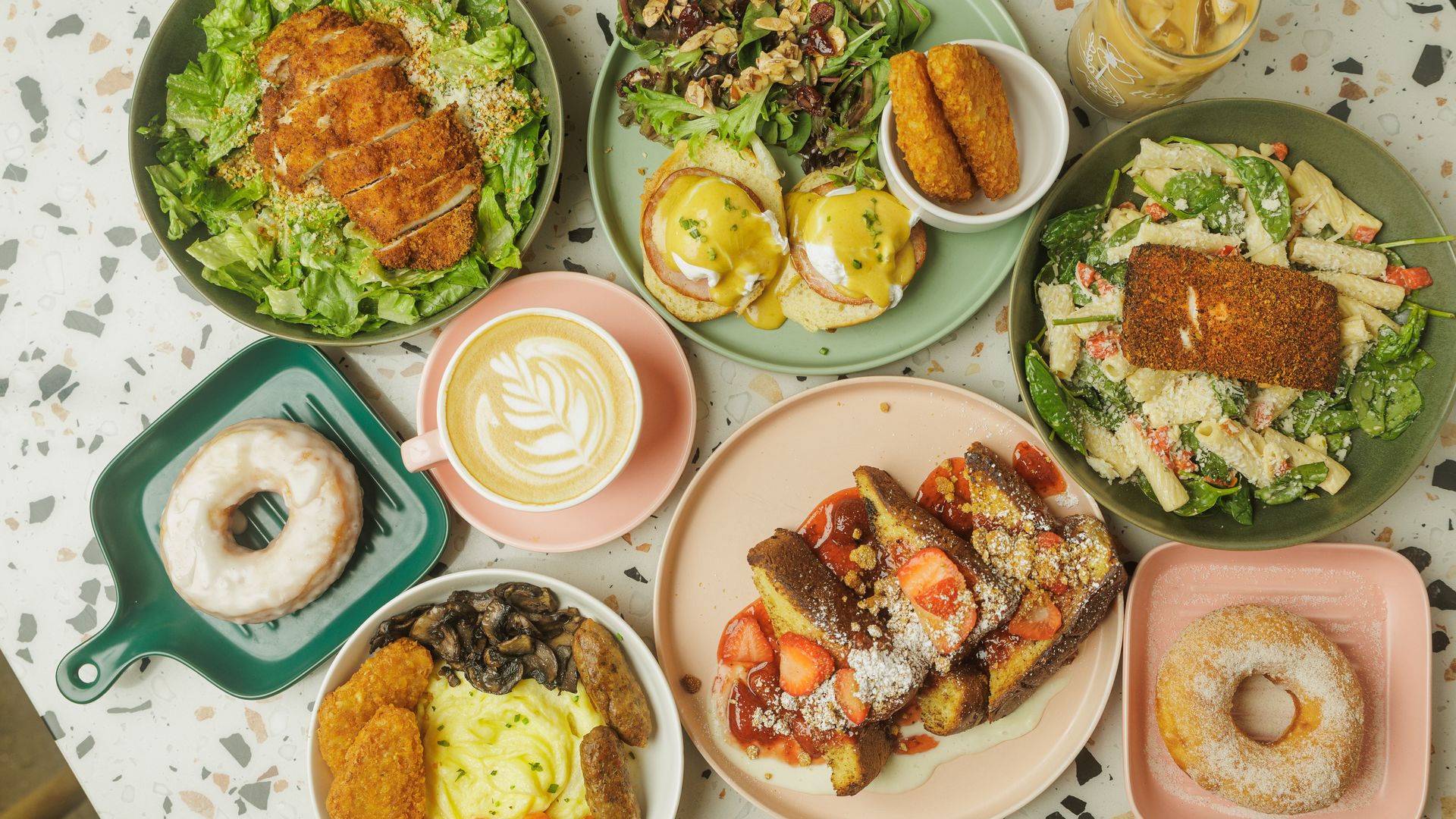 An overhead spread of colorful plates: crispy breaded chicken on greens, a greens salad with nuts, Eggs Benedict with hollandaise, pasta with chicken, donuts, iced coffee, and various sides.