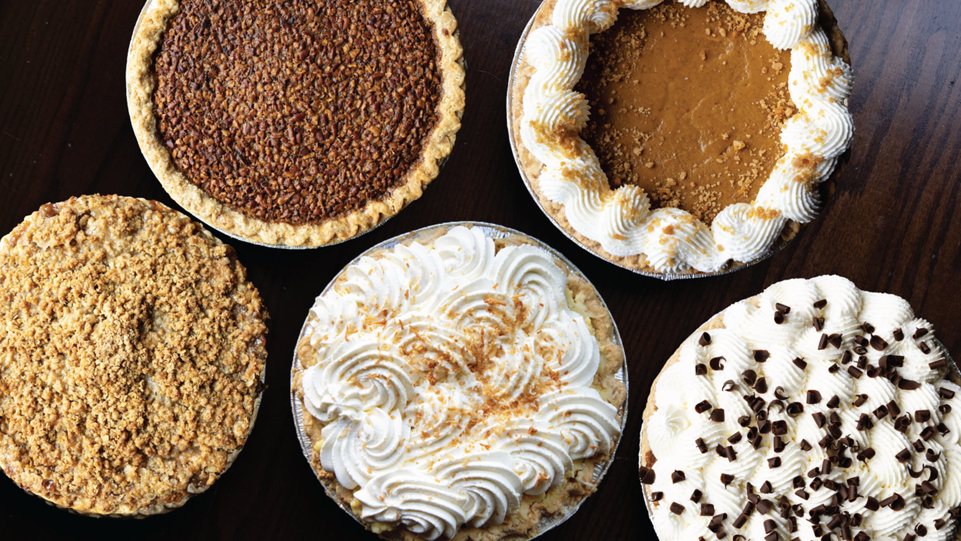 Five assorted pies on a dark wooden table, including pecan, pumpkin with whipped cream, crumb-topped, whipped cream with crumbs, and whipped cream with chocolate shavings.