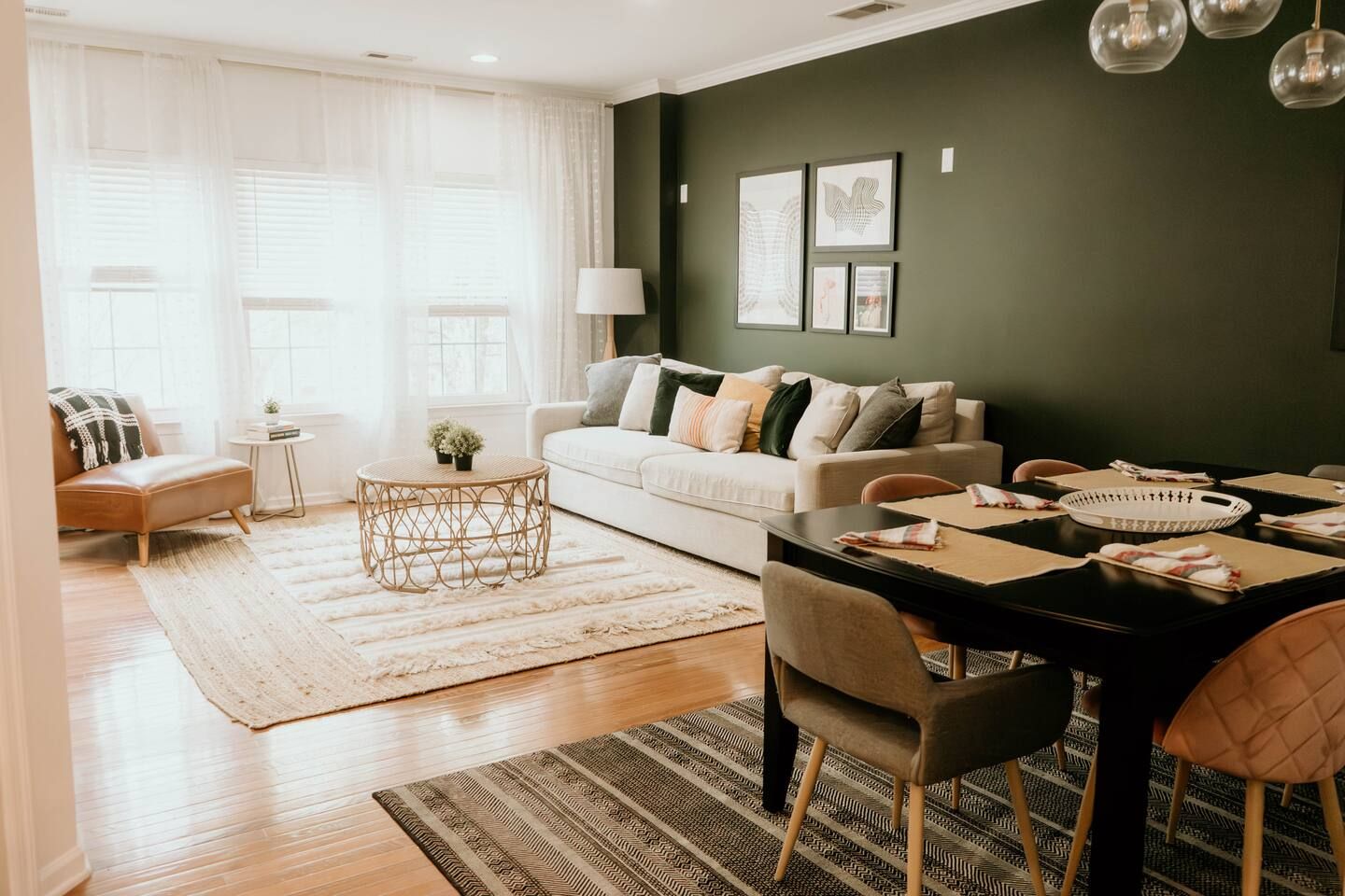 Bright living room with large windows, sheer white curtains, tan leather chair with black plaid blanket, beige sofa with various pillows, round rattan coffee table, dark green accent wall with four framed artworks, and black dining table with mixed chairs and place settings.