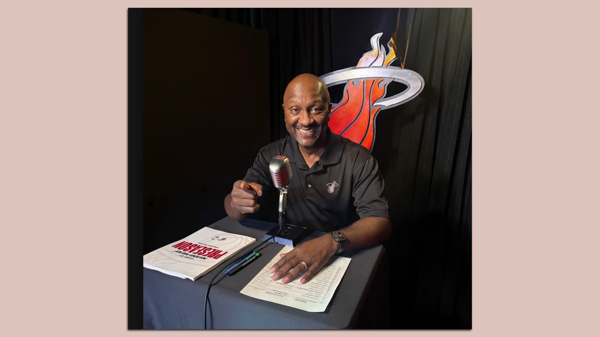 Smiling bald man in a black polo sits at a table with a vintage microphone, pointing at the camera. A flaming basketball logo is behind him; papers and pens on the table.