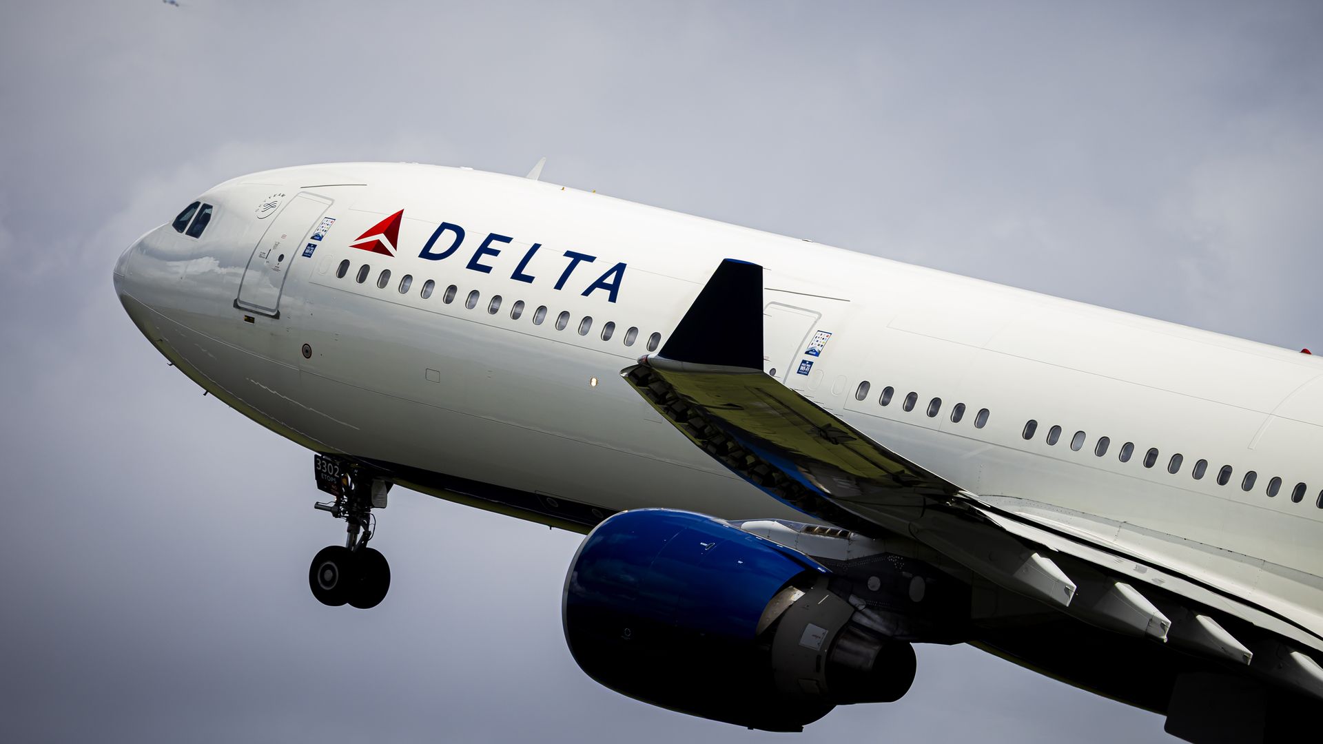 Delta Air Lines jet ascending against a cloudy sky; white fuselage with blue Delta lettering and red logo, blue engine, nose gear down, wing flaps extended.