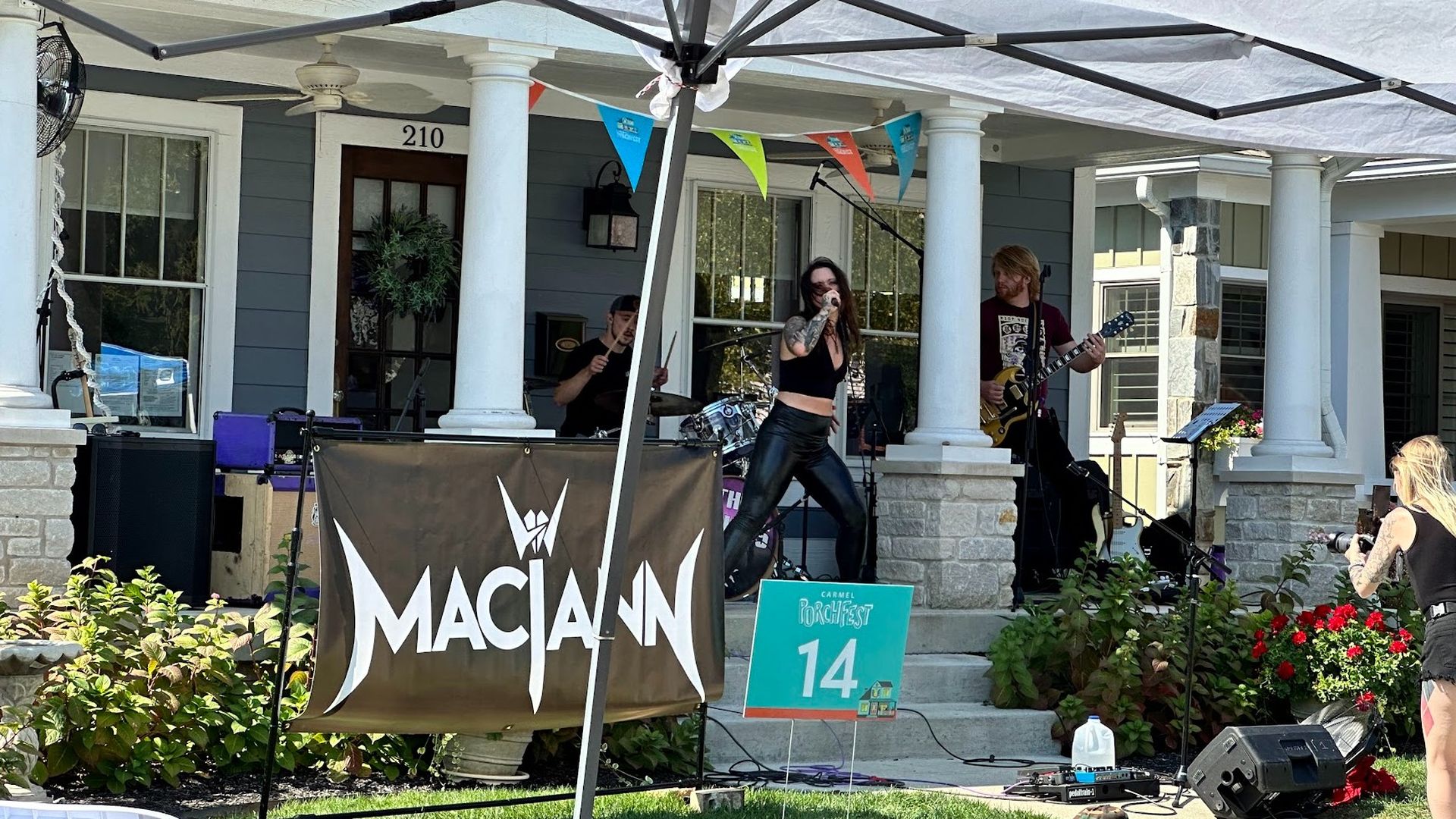 Outdoor live music performance on front porch of house number 210 with three band members playing, colorful flags, a banner reading "MACJANN," and a sign for Carmel Porchfest 14.
