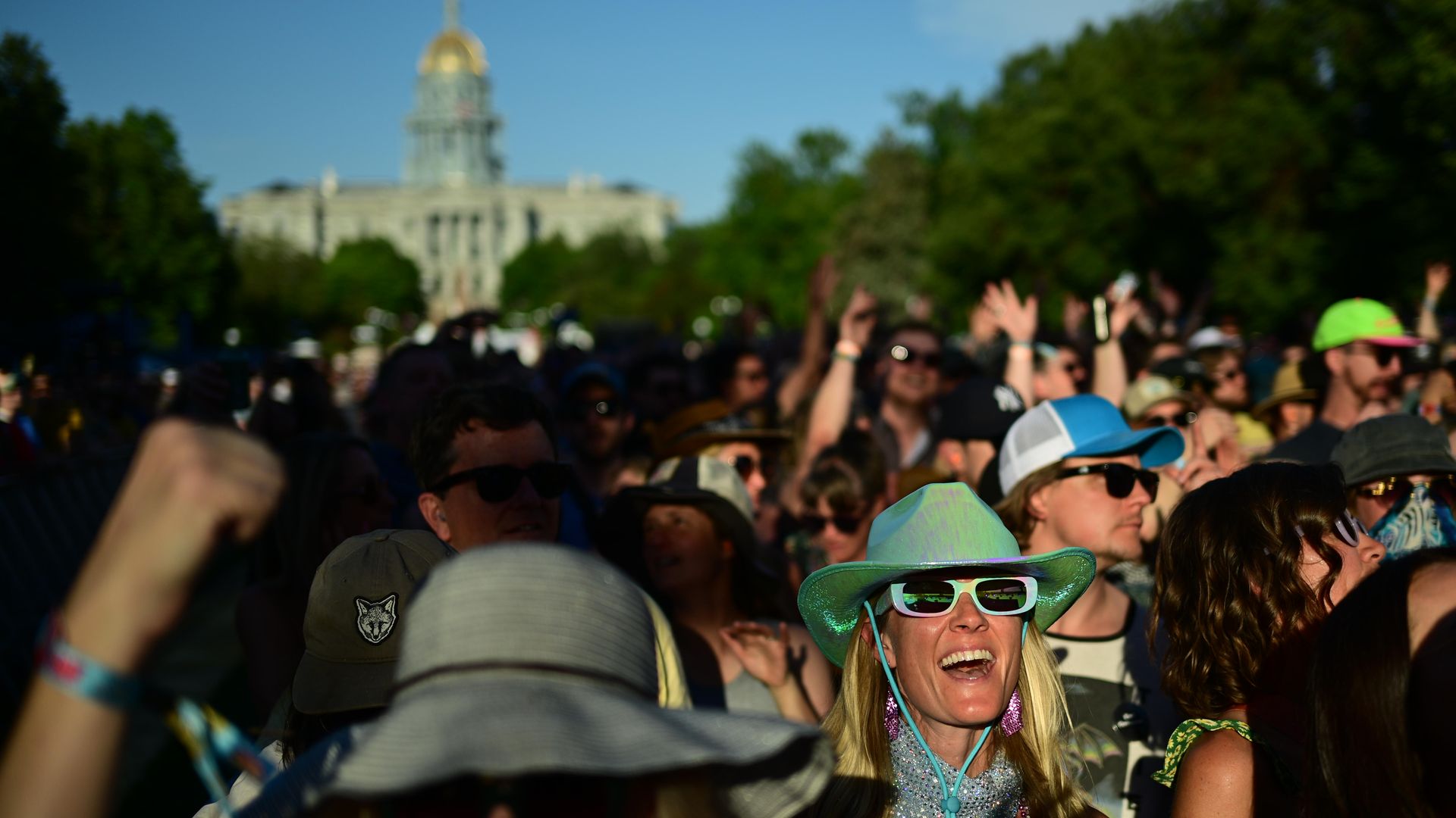 Crowd at an outdoor event with many people wearing sunglasses and hats; a smiling woman in a bright green hat and white sunglasses in the foreground. Trees and a domed building blur in the distance.