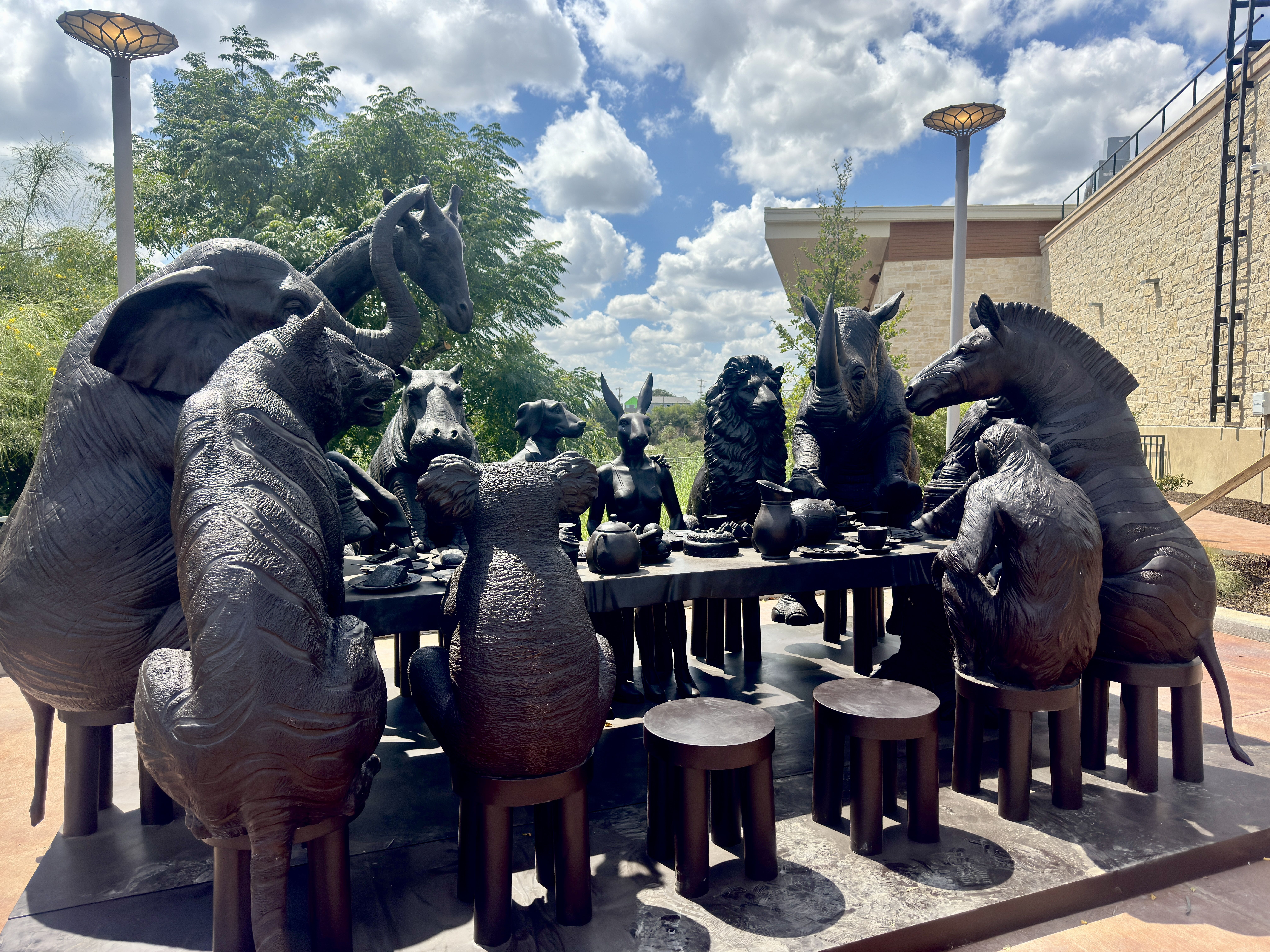 Bronze statue of various animals including elephant, rhino, zebra, lion, giraffe, and more, seated around a table having tea outdoors under a partly cloudy sky.