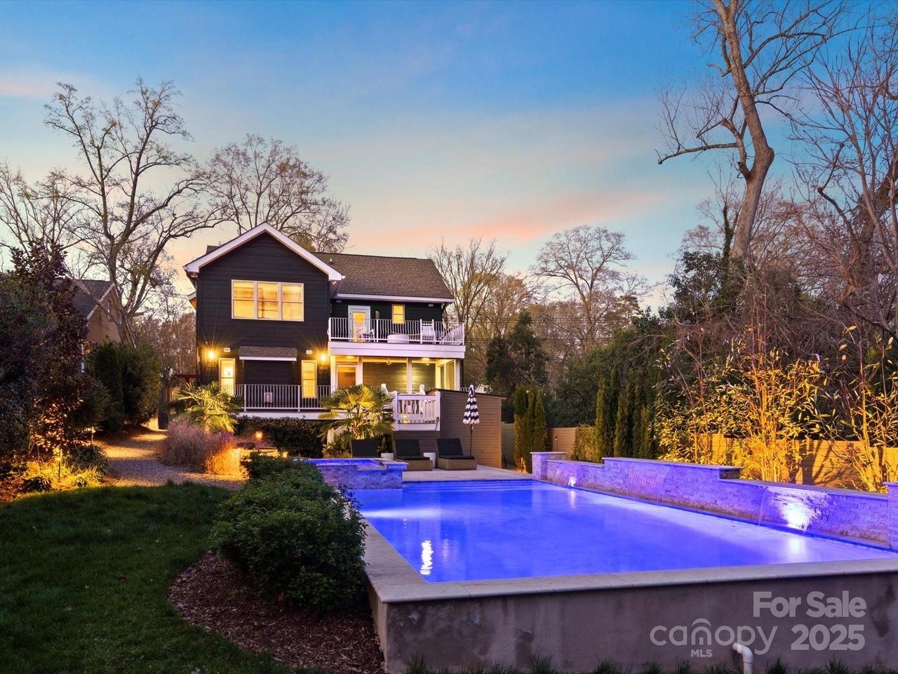 Twilight view of a black two-story house with lit windows, white balcony railings, and a glowing blue pool in the foreground, surrounded by trees and outdoor seating.