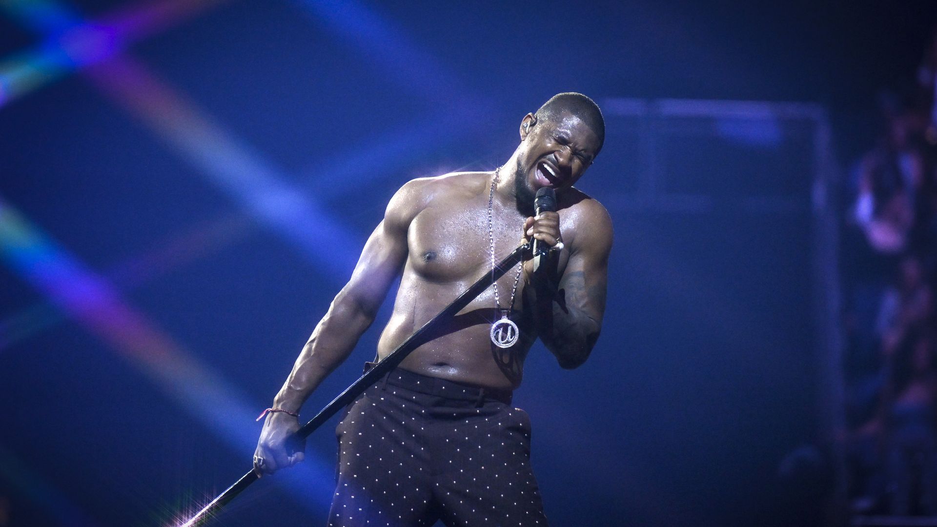 A shirtless male performer on stage, sweating and singing into a microphone, wearing black studded pants and a silver chain, under blue stage lights with colorful beams in the background.