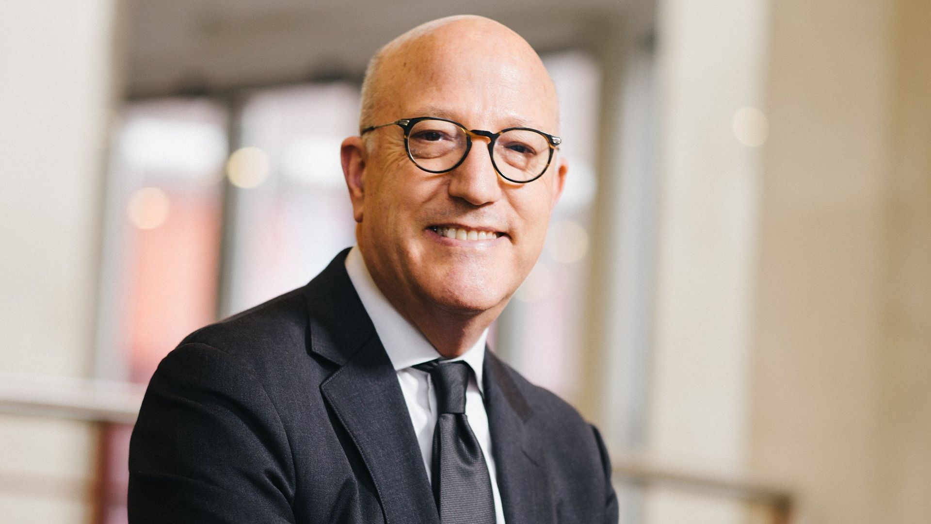 Smiling bald man in a dark suit and round glasses sits indoors, with a soft blurred background of a railing and windows.