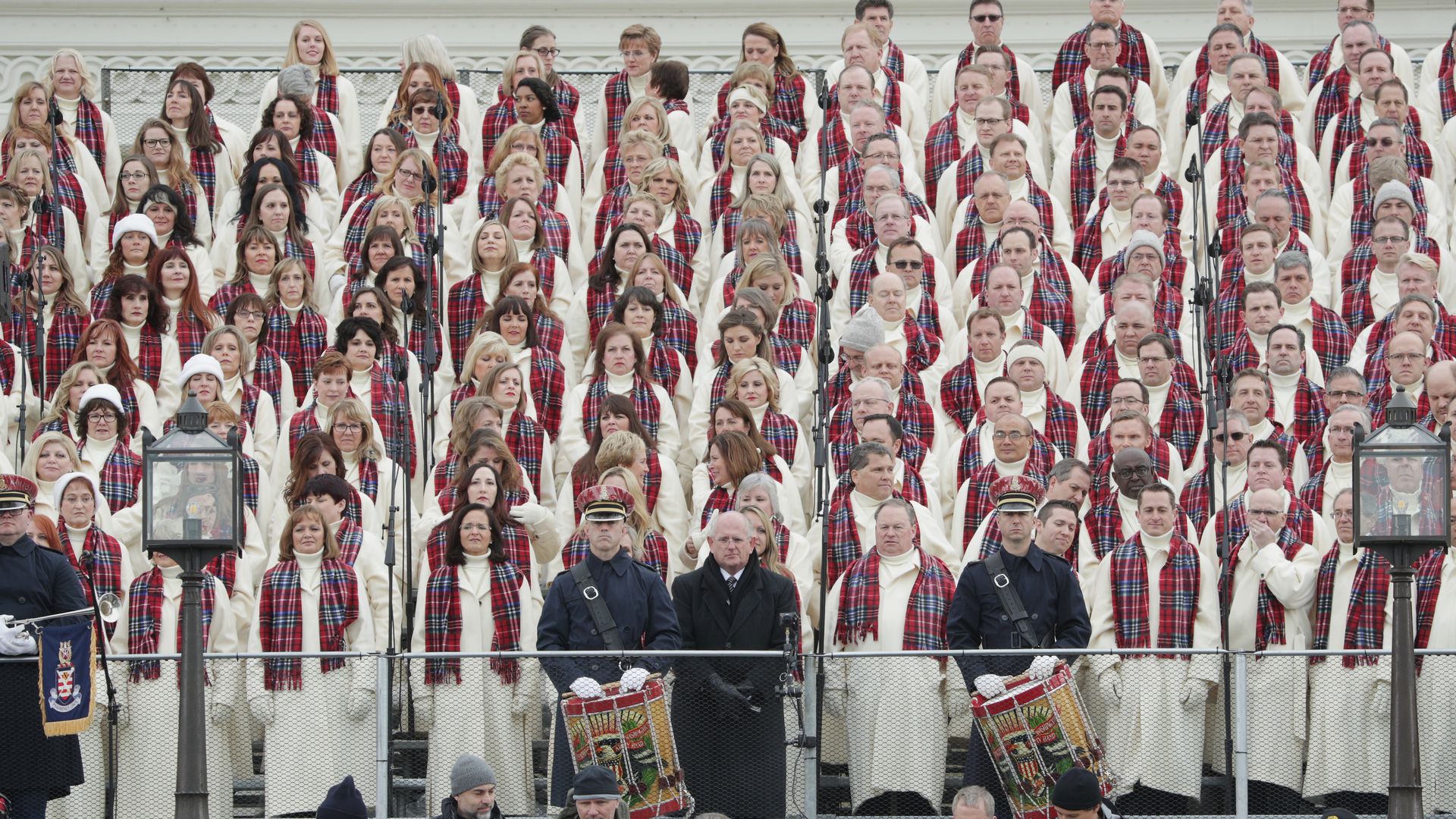 A choir in white robes and red plaid scarves behind two military officers and a man in a suit.