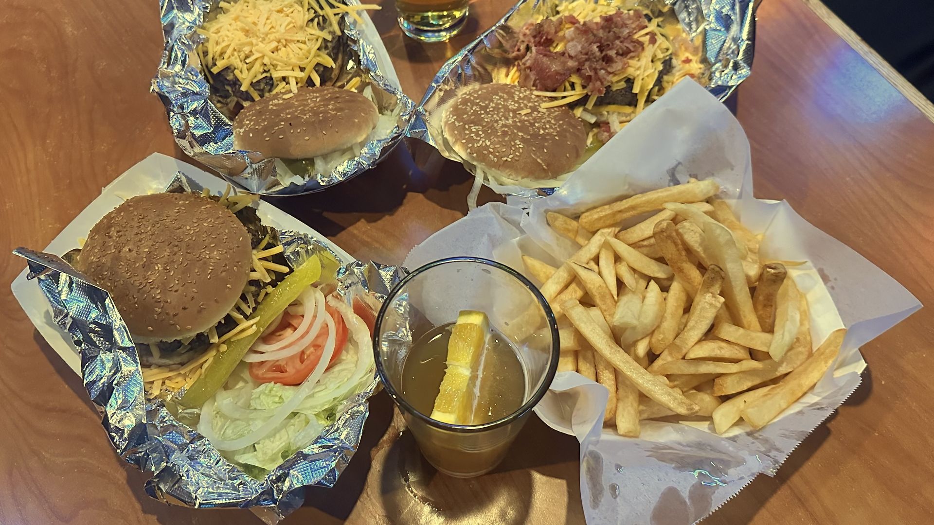 Three cheeseburgers with onions, lettuce, tomato, pickles, and bacon on foil-lined trays, two drinks including one with a lemon slice, and a basket of French fries on a wooden table.