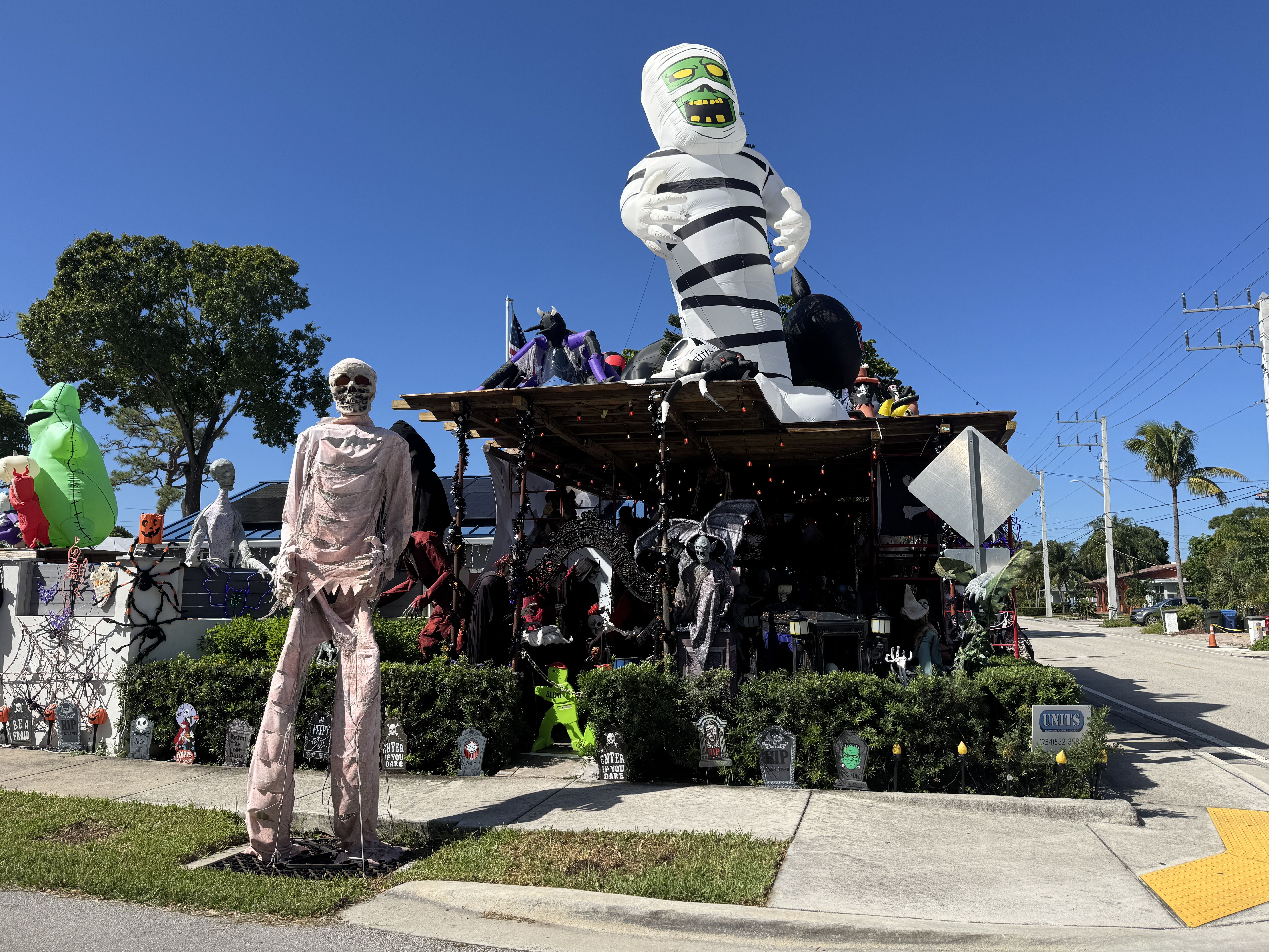 Bright Halloween float features a towering white and black striped inflatable mummy with a green face and yellow eyes, surrounded by skeletons, tombstones, and cobwebs against a blue sky.