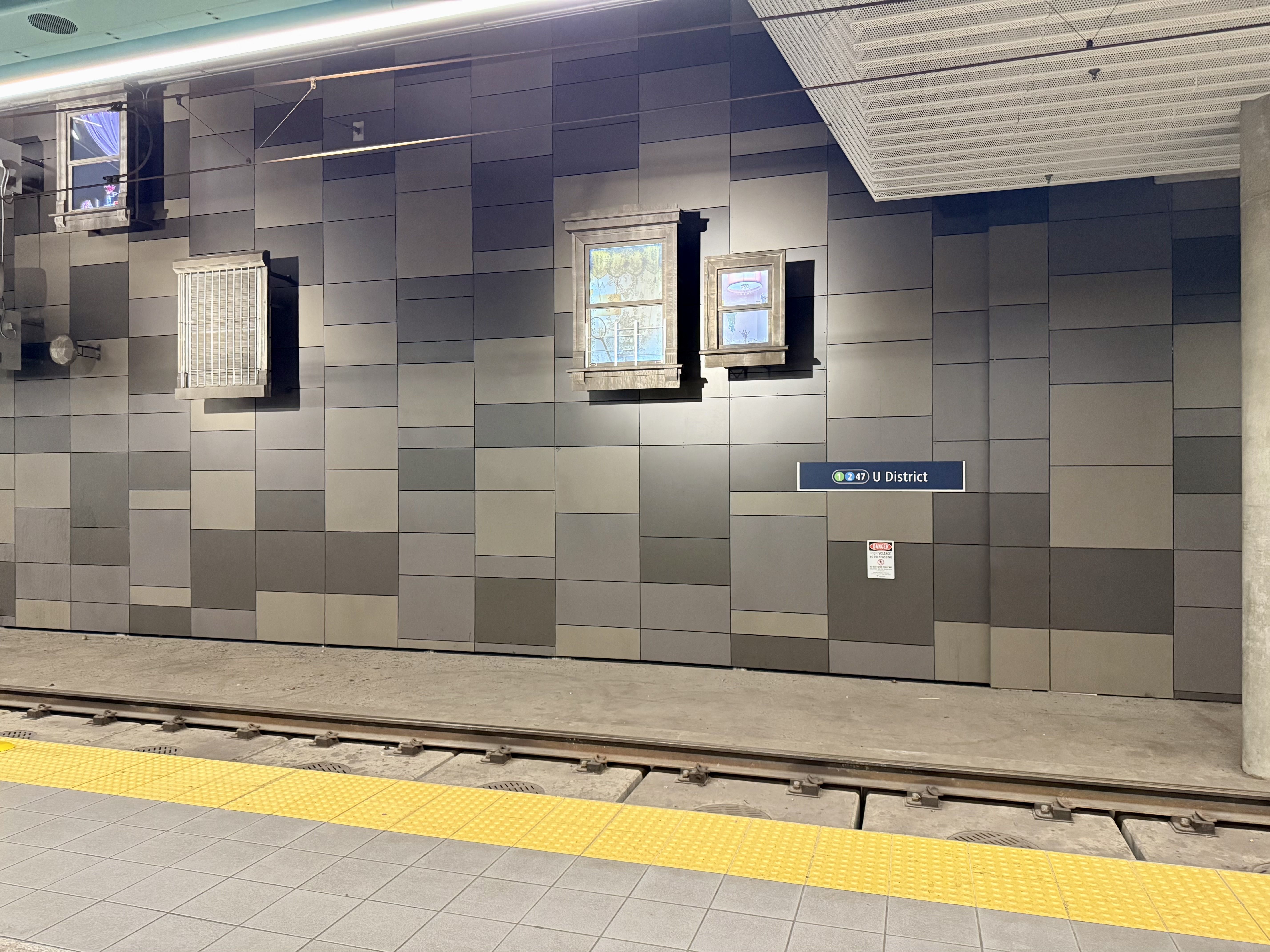 Underground light rail station platform with a grey tiled wall, two small windows, a bright yellow tactile edge strip, and a blue "U District" sign near the tracks.