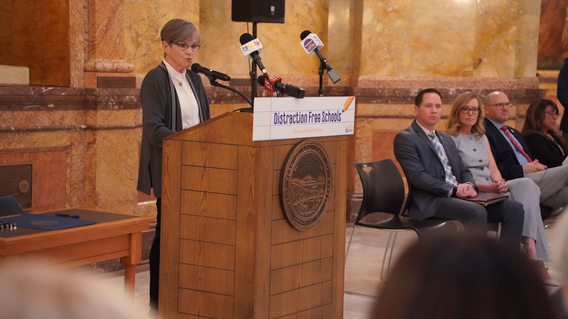 An older woman with short gray hair and glasses speaks at a wooden podium in a marble chamber; a "Distraction Free Schools" sign hangs on the front as suited colleagues sit behind her.