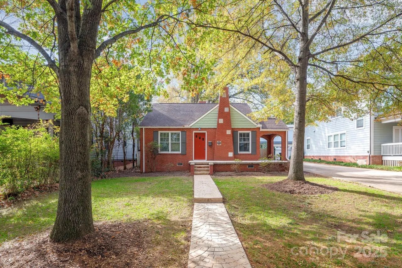 Small red-brick house with a red door, front chimney, and green siding at gable, with a concrete path leading to the door, flanked by two large trees in a grassy front yard.