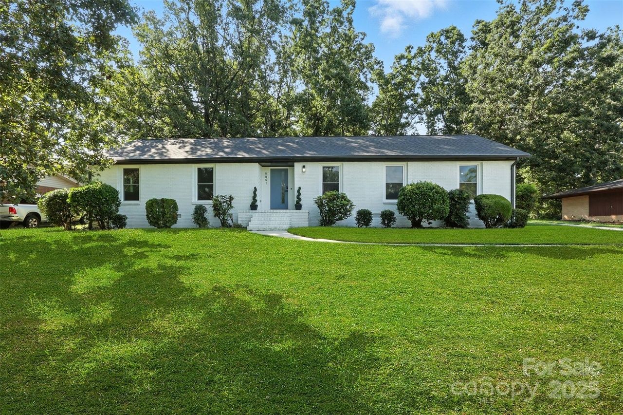 Single-story white brick house with gray roof, blue front door, surrounded by green bushes and trees, with a large green lawn and a curved pathway leading to the entrance, under a blue sky.