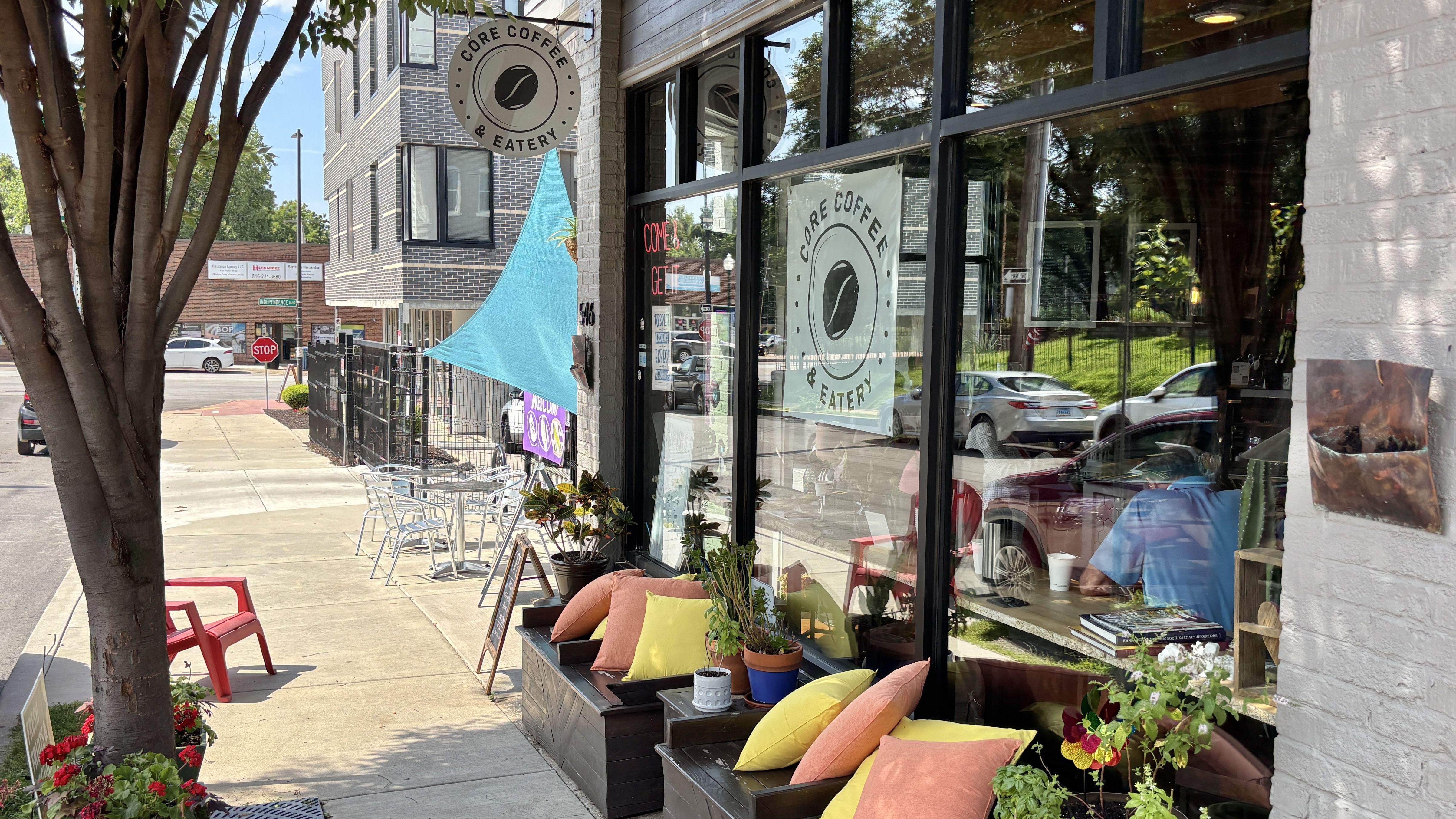 Exterior of Core Coffee & Eatery: circular logo sign, large storefront windows with a poster, and a colorful sidewalk seating area with cushions; blue shade sail, trees, and passing cars.