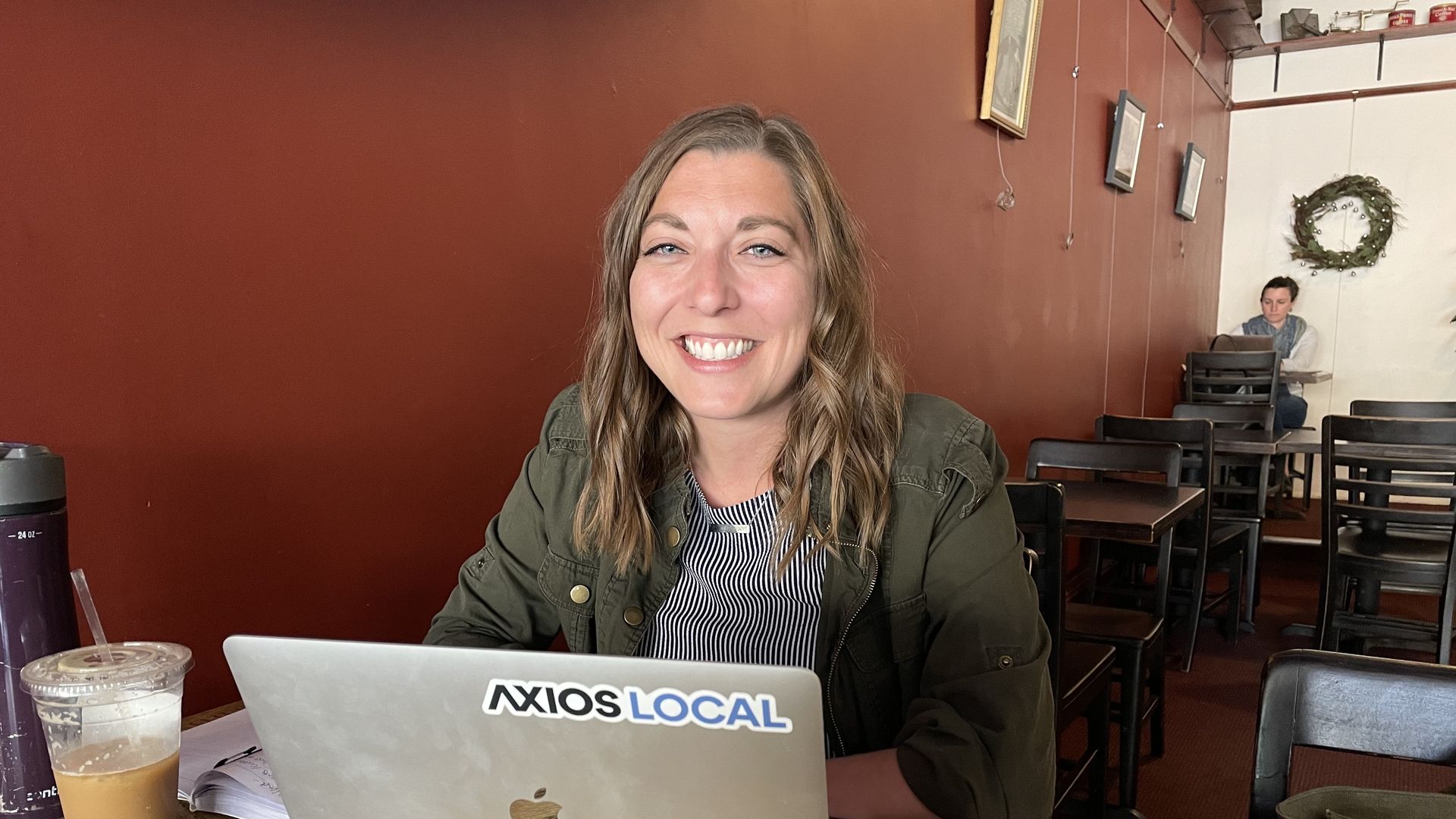 Smiling woman with wavy brown hair in green jacket working on a laptop with an "AXIOS LOCAL" sticker in a café with red walls, wooden tables, drinks, and another person in the background.