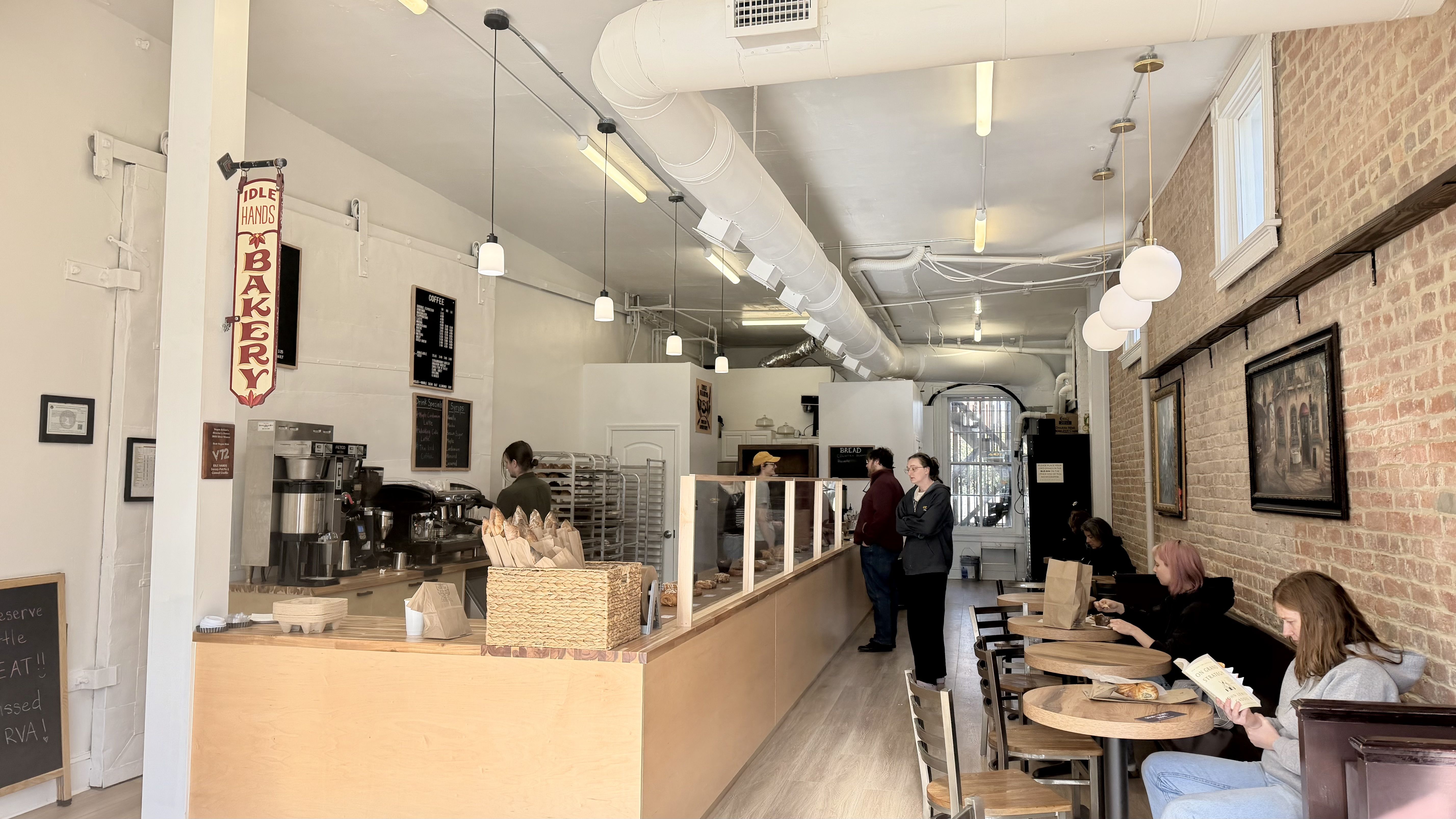 Bright bakery cafe with a counter of bread and pastries, espresso station, and a hanging "Idle Hands Bakery" sign. White walls, exposed ducts, brick wall with lights and customers at tables.