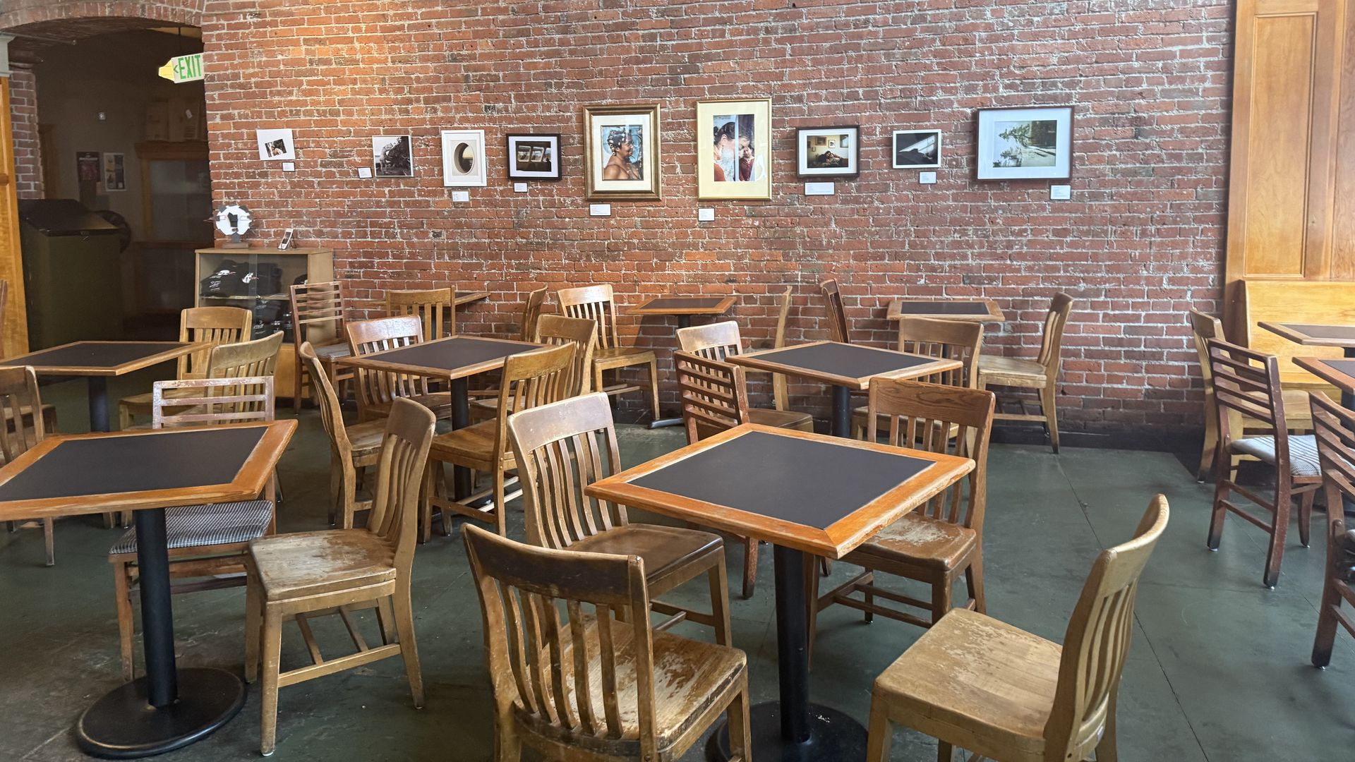 Empty cafe with wooden chairs and tables, a brick wall backdrop featuring eight framed art pieces, and an exit sign above a doorway in the left corner.