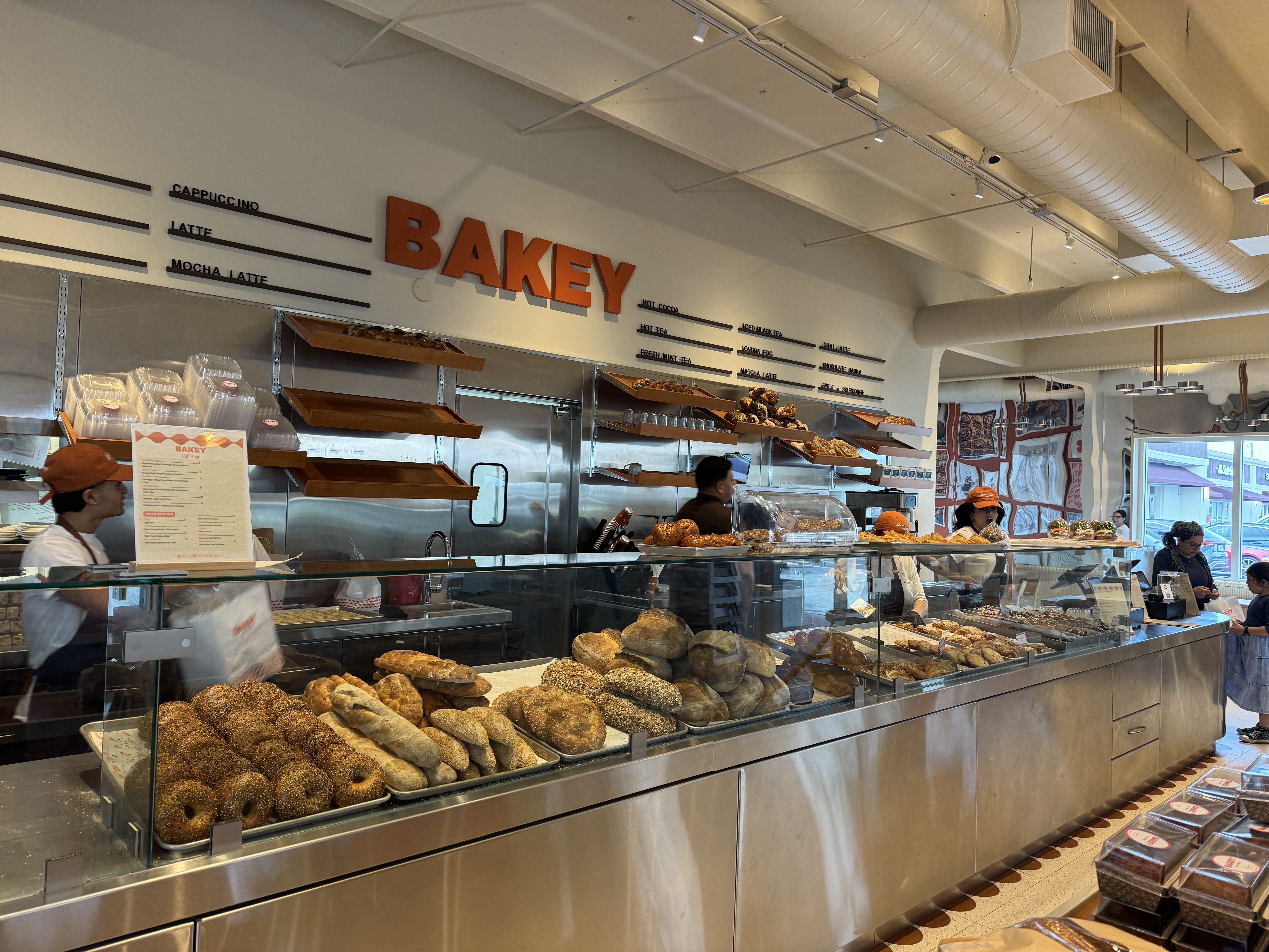 Inside a bakery shop with a glass display case of sesame bagels and loaves. A large orange sign reads "BAKEY" on the wall; staff in orange caps work behind the counter under exposed ducts.