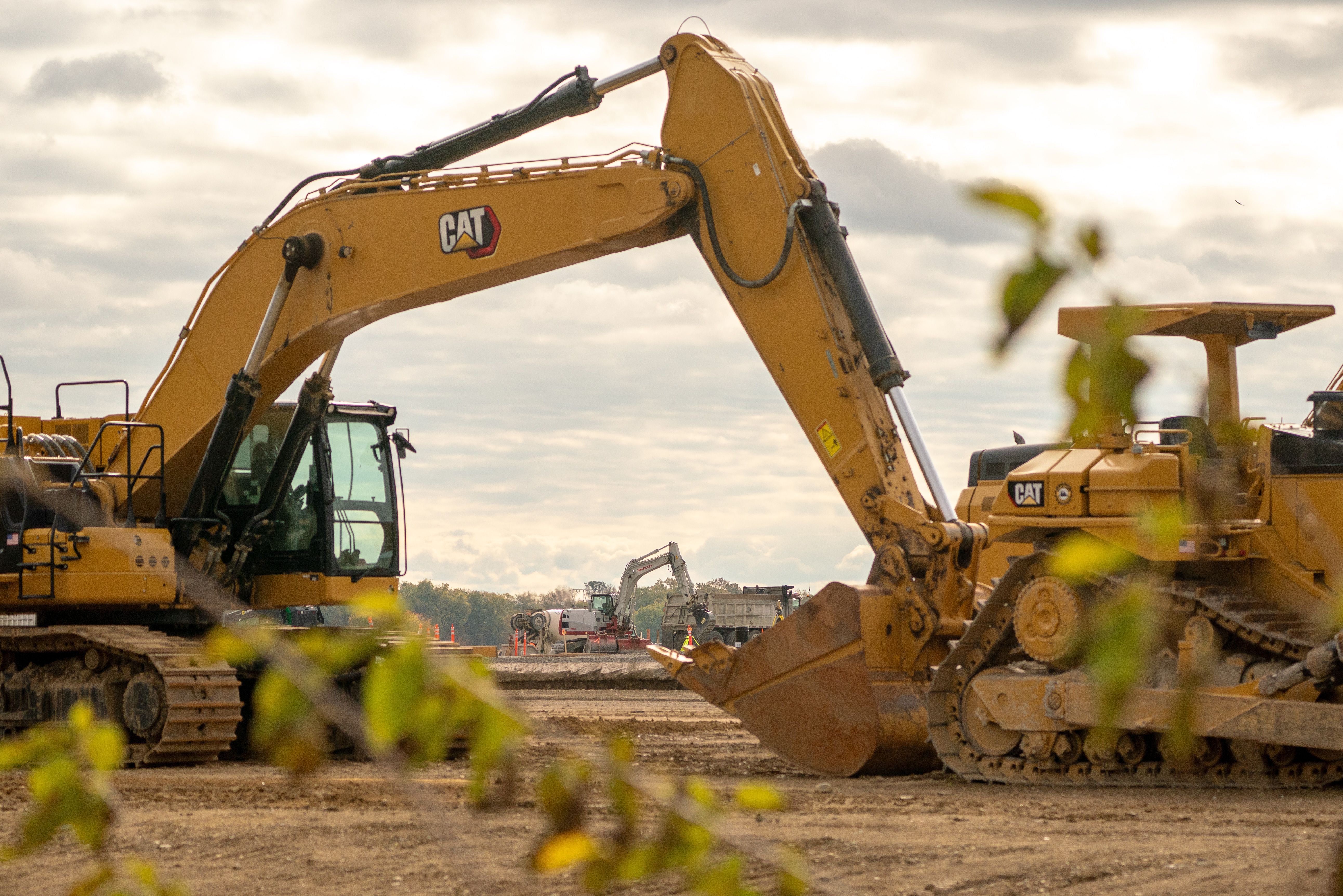Yellow CAT excavator and bulldozer at a construction site with dirt ground and cloudy sky, some green leaves in foreground, other machinery and workers in background.