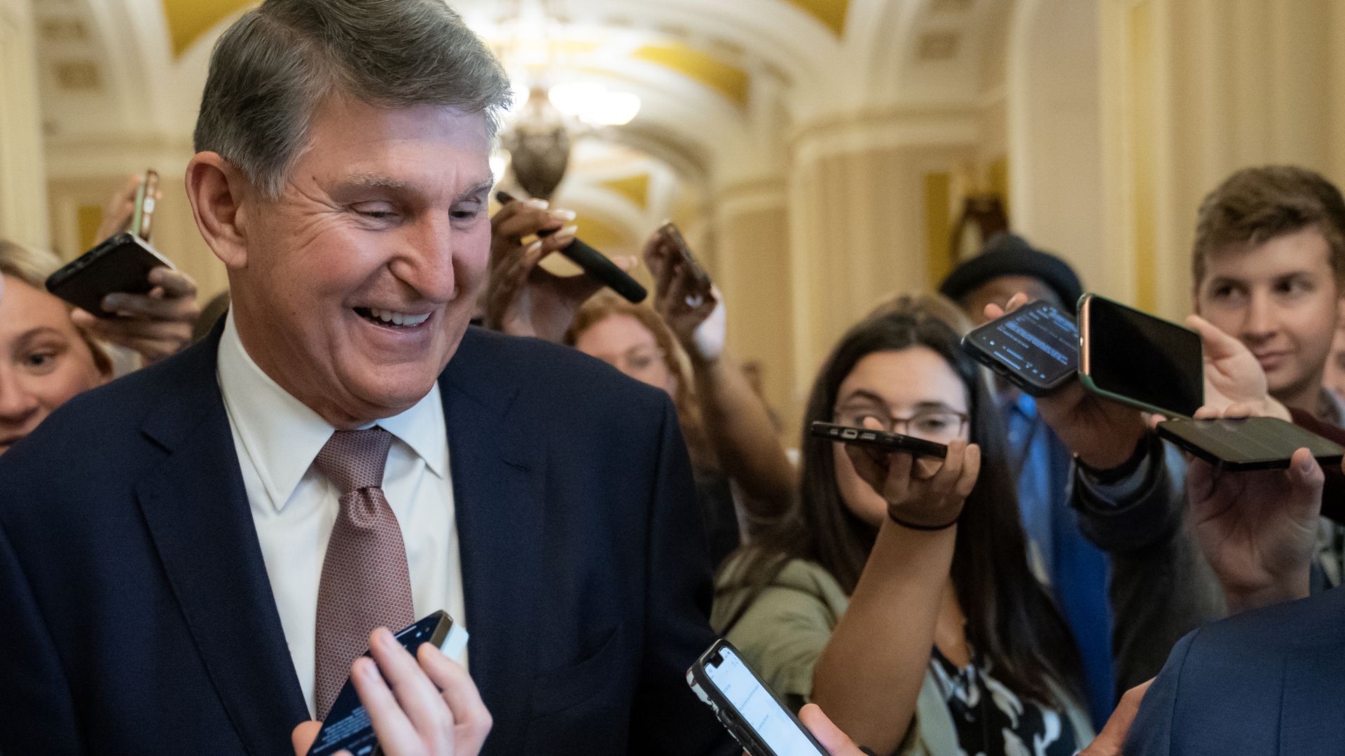 Sen. Joe Manchin, wearing a dark suit, white shirt and brown tie, talks with reporters.