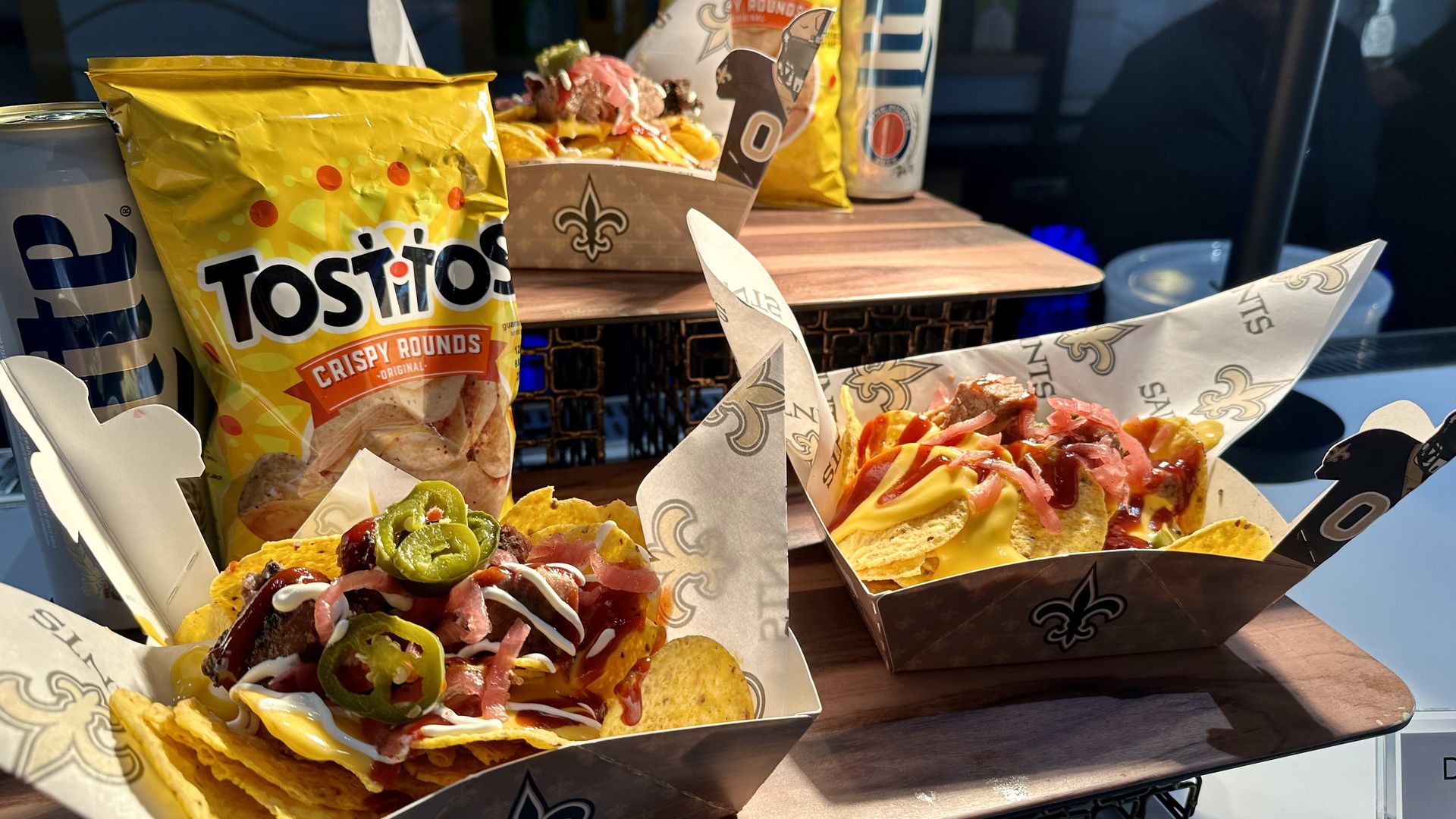 Two trays of loaded Tostitos nachos topped with jalapeños, cheese, pickled onions, and sauces, with a yellow Tostitos bag and a can of Miller Lite beer in the background.