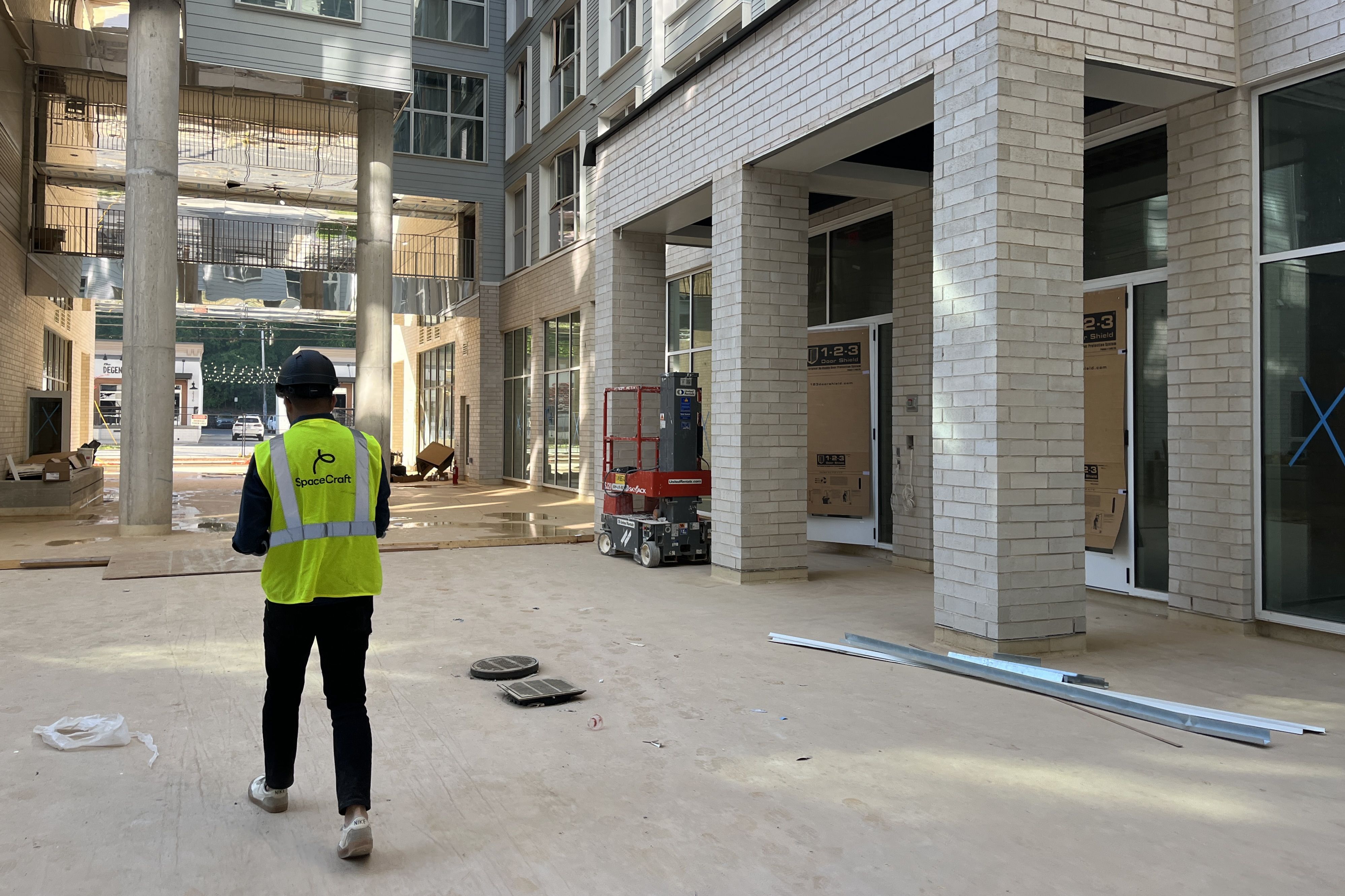 Construction worker in a neon yellow safety vest and hard hat walks through a modern courtyard under construction, with brick columns, glass storefronts, and scaffolding above.