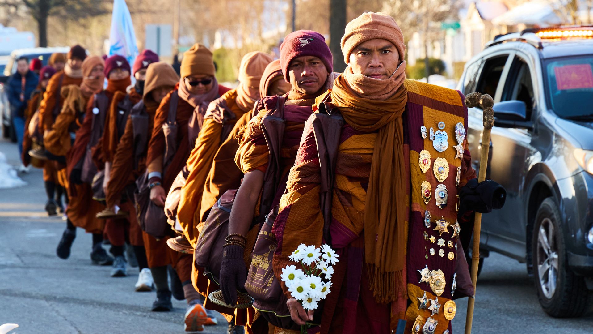 Monks in the "Walk for Peace" dressed in brown and maroon robes walk outdoors in cold weather, one holding white flowers and a wooden staff, with a car nearby and snow on the ground.