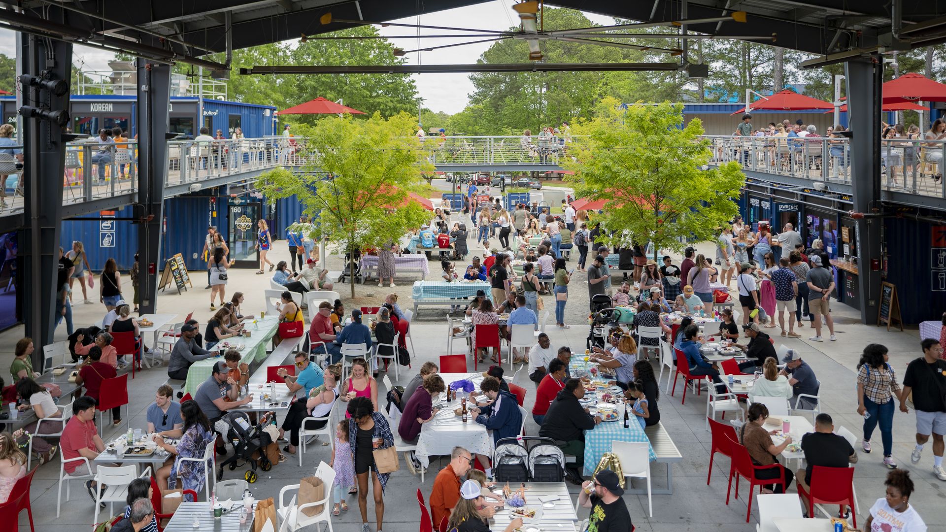 Busy outdoor food court with people eating, walking, and socializing. Tables have colorful checkered tablecloths. Blue food containers line the sides, and green trees and red umbrellas add color.