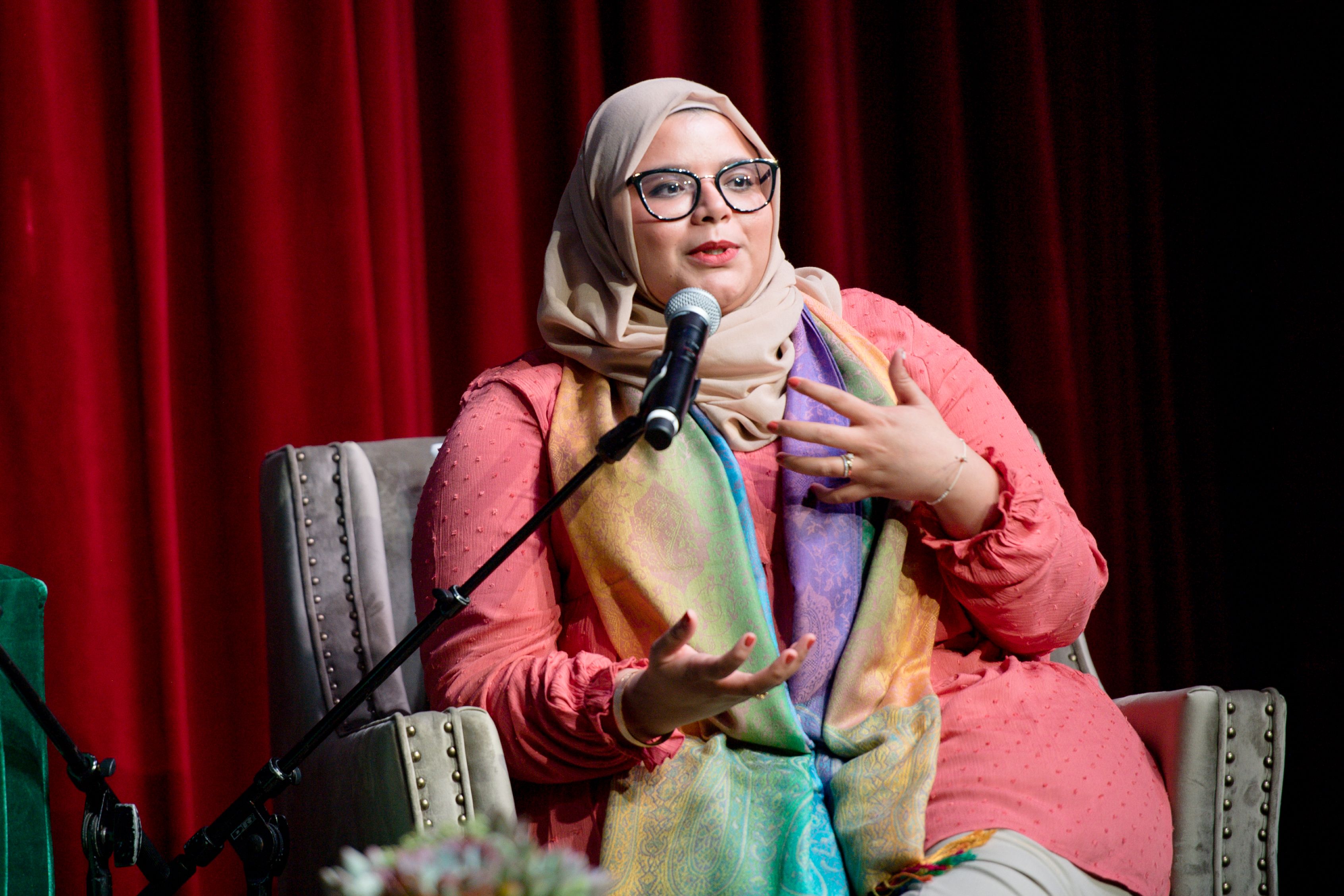 A woman wearing a hijab and glasses speaks into a microphone while sitting on a stage
