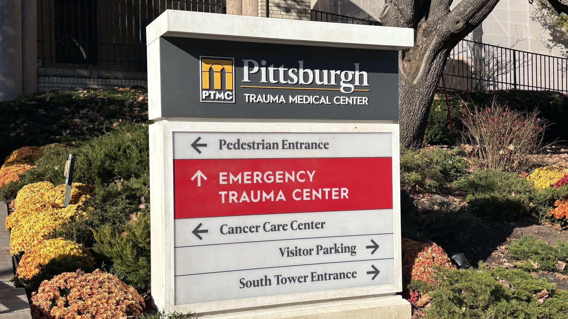 Sign for the fake "Pittsburgh Trauma Medical Center" with PTMC logo; white and red panels list directions (Pedestrian Entrance, Emergency/Trauma Center, Cancer Care, Visitor Parking) beside autumn shrubs.