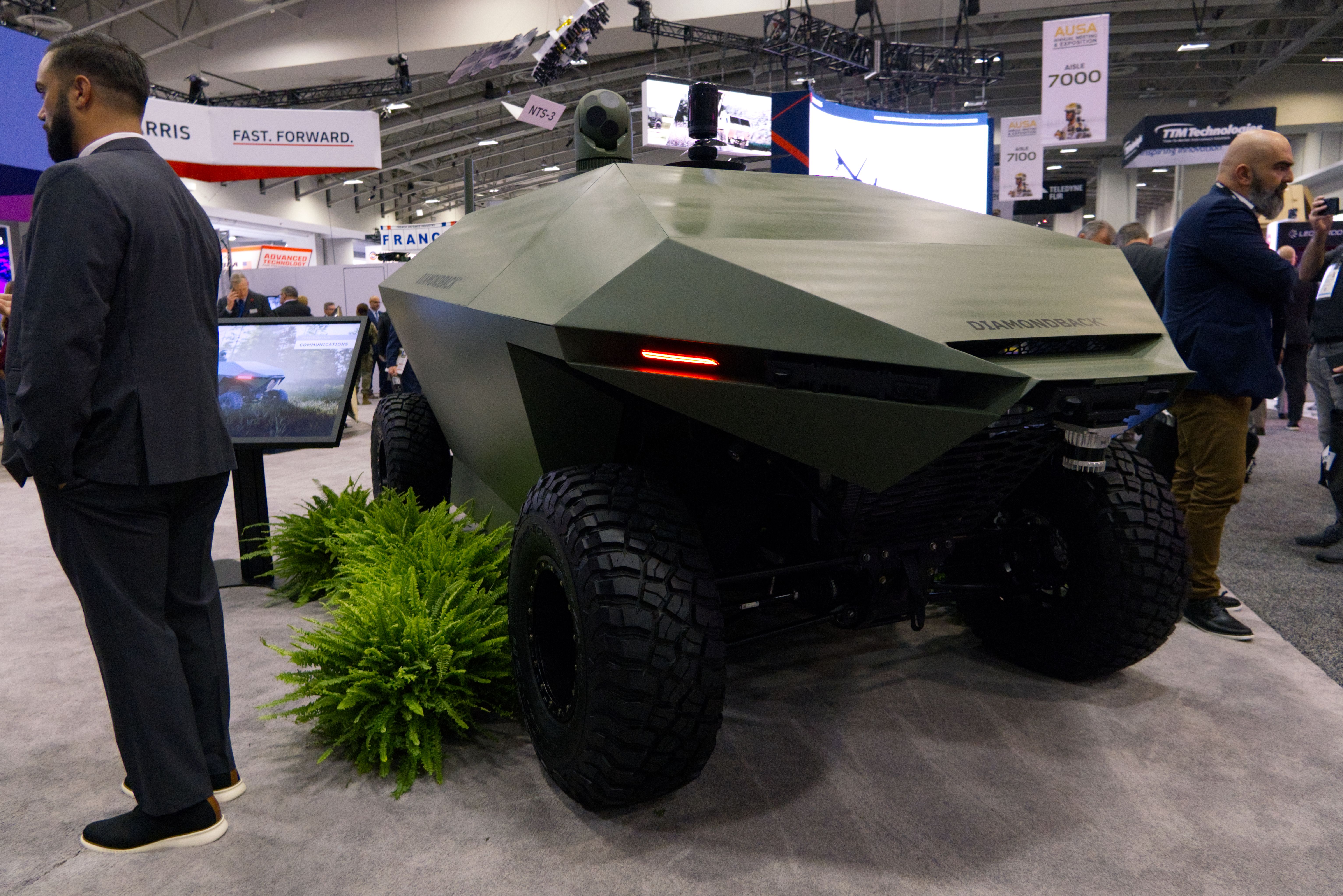 An unmanned ground vehicle is parked on the AUSA show floor. The carpet is grey. It is near two fake ferns.