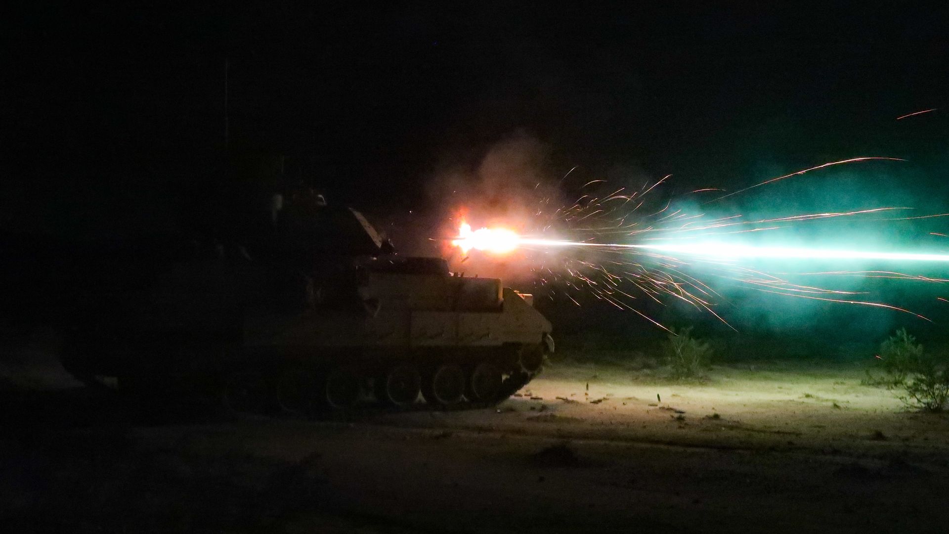 A dark nighttime scene with a large combat vehicle shooting its main gun.