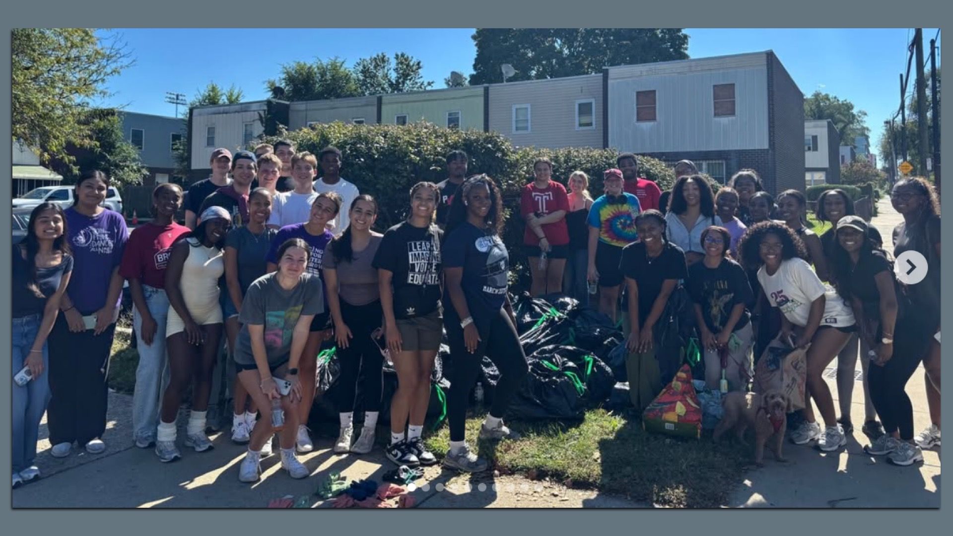 Large diverse group of volunteers poses outdoors on a sunny sidewalk in front of a row of houses, smiling at the camera, with piles of gathered bags at their feet.