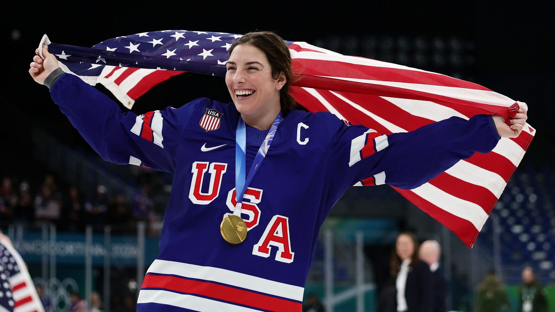 Hilary Knight wears her red, white and blue Team USA uniform and her new gold medal while holding an American flag over her shoulders that billows behind her. The arena crowds are visible in the stands behind her. 