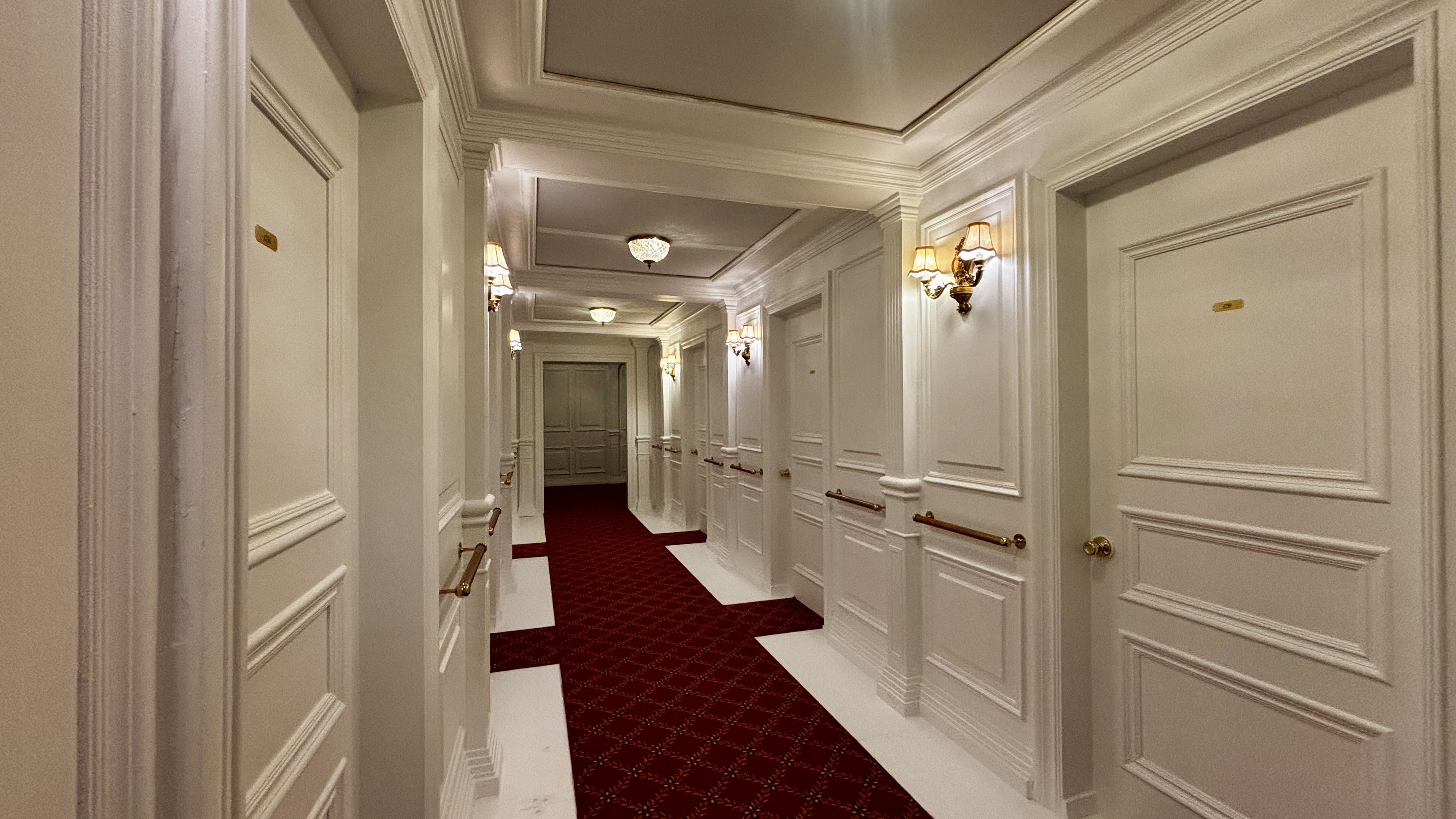 Narrow hotel hallway with white paneled walls, red patterned carpet, multiple white doors with gold handles, brass handrails, and warm wall sconces lighting the space.
